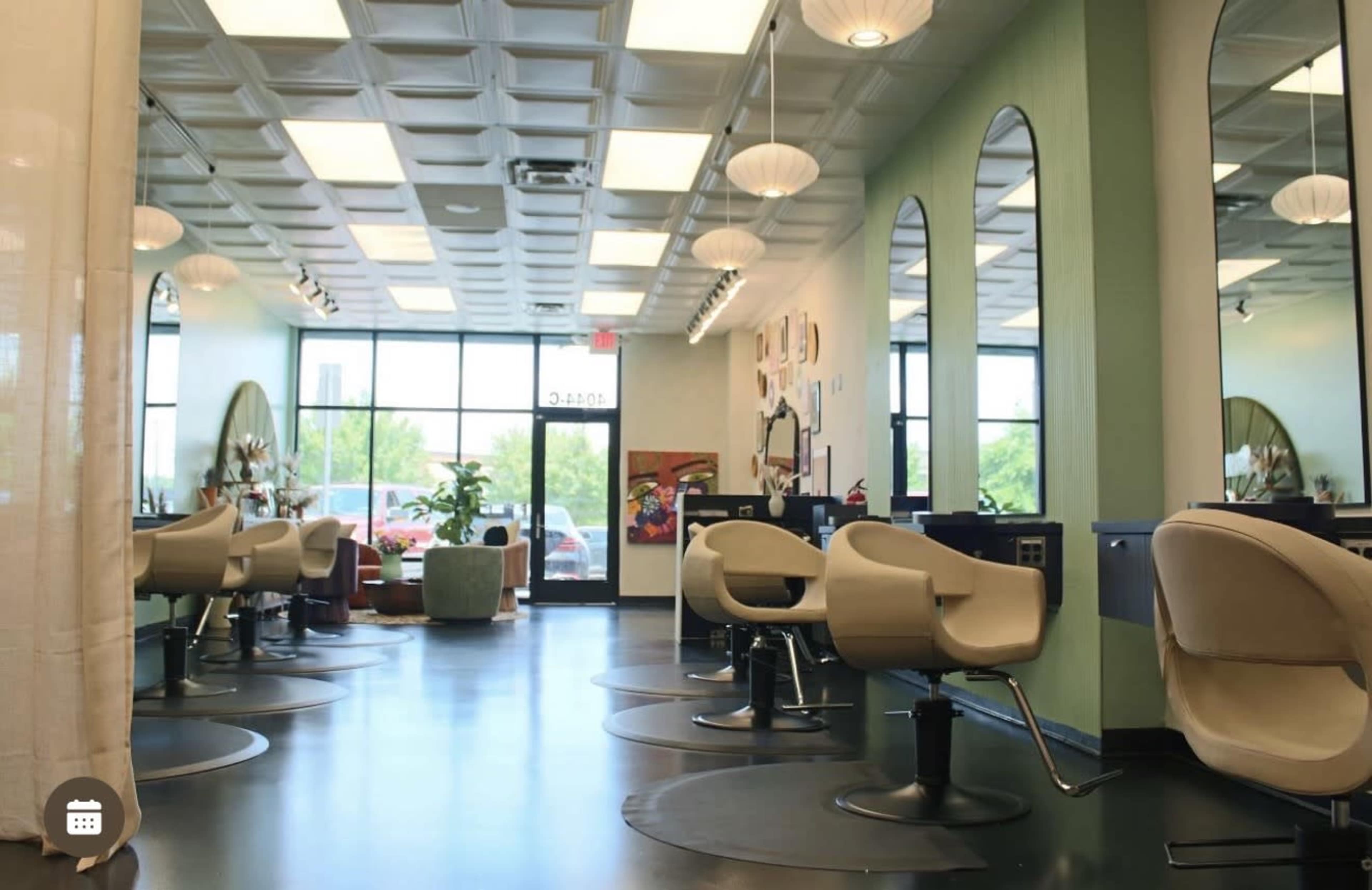 The interior of a salon features several styling chairs and large mirrors, with natural light streaming in through large windows.