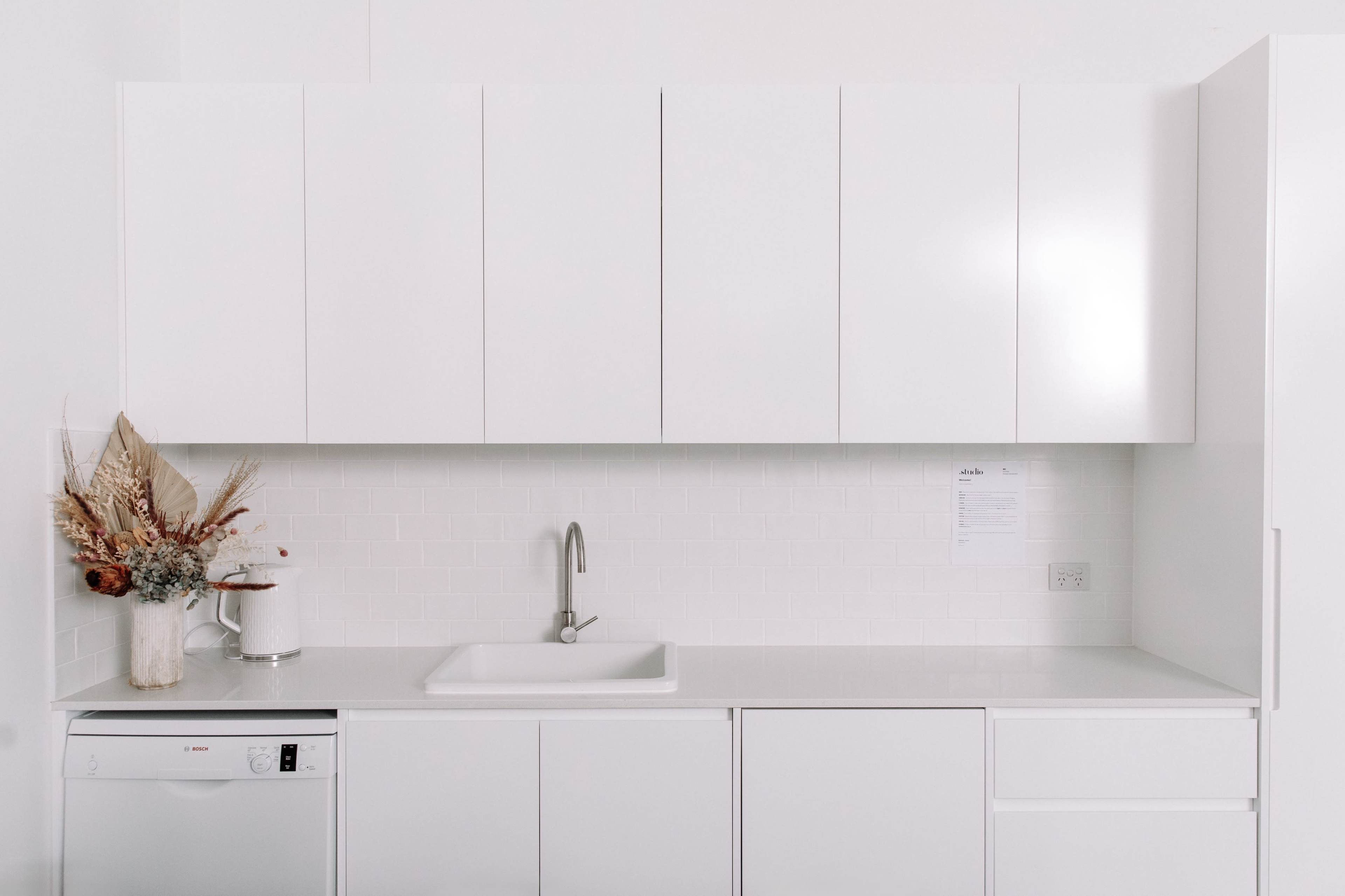 The image shows a minimalist kitchen with white cabinetry, a stainless steel sink, and a small decorative arrangement of dried flowers on the counter.
