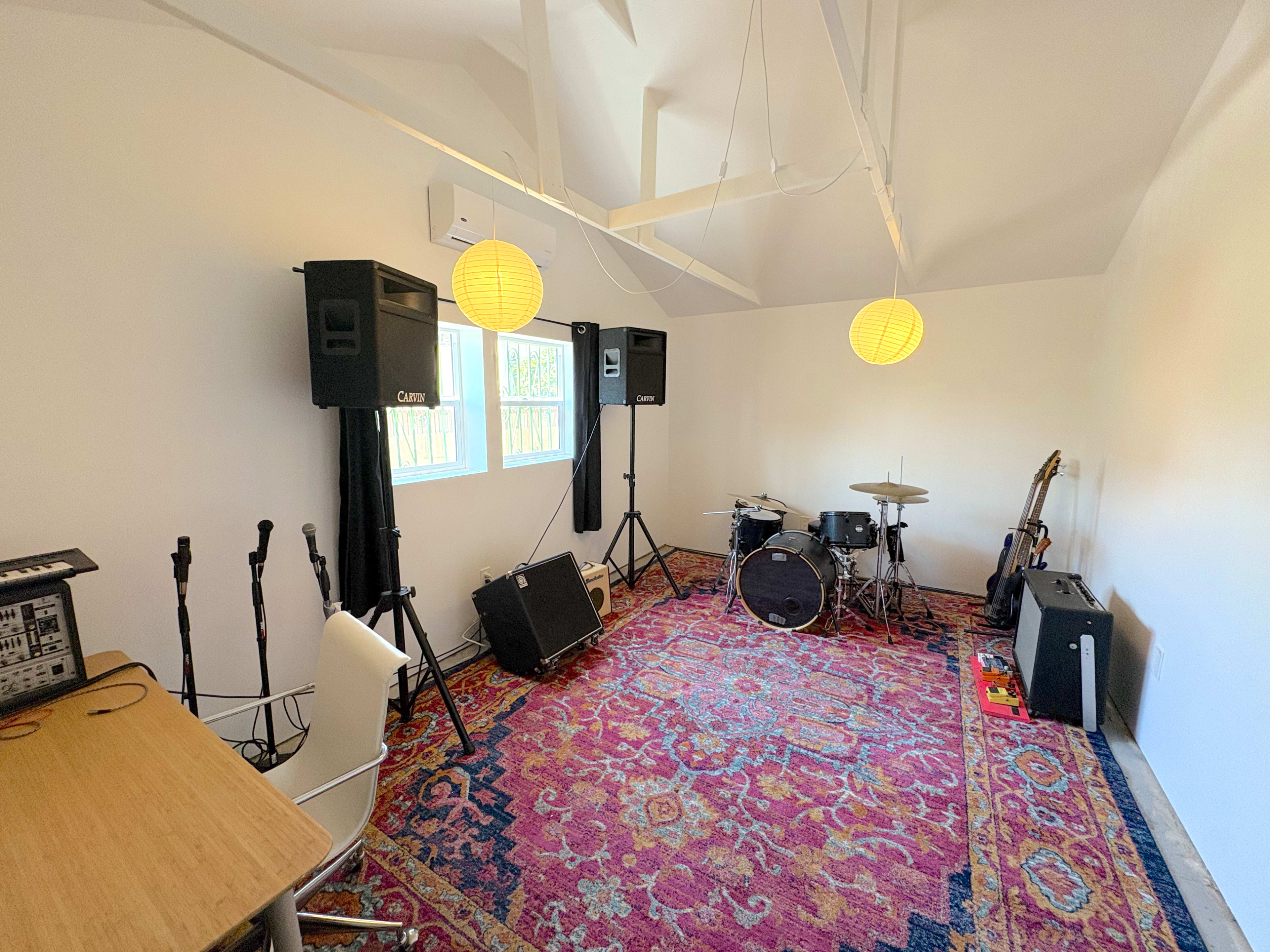 The room features a music setup with a drum kit, amplifiers, microphones, and a desk, all arranged on a patterned rug.