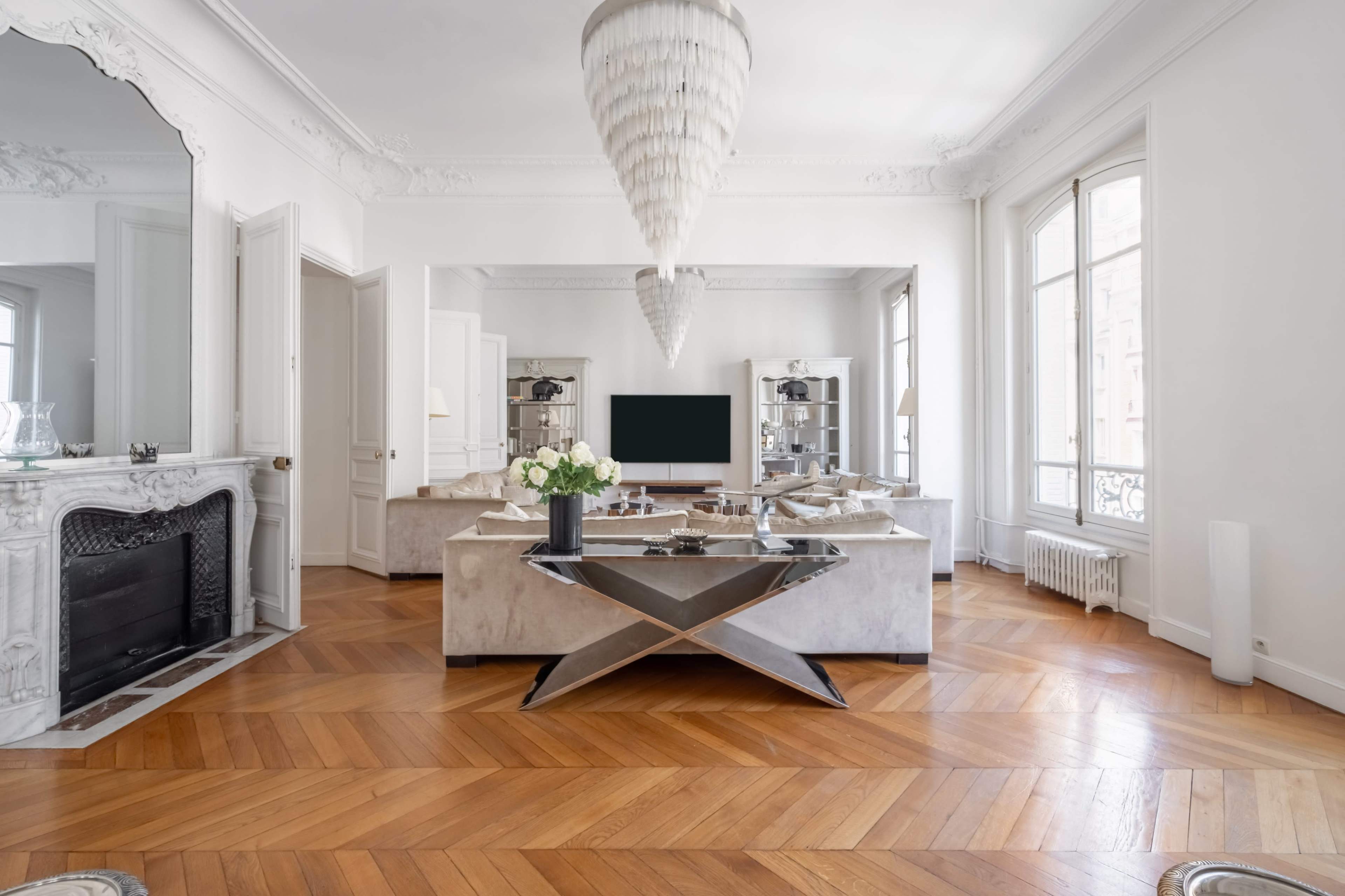 The image shows a modern living room with a decorative chandelier, hardwood flooring, a fireplace, and a large television positioned within a stylish arrangement of furniture.