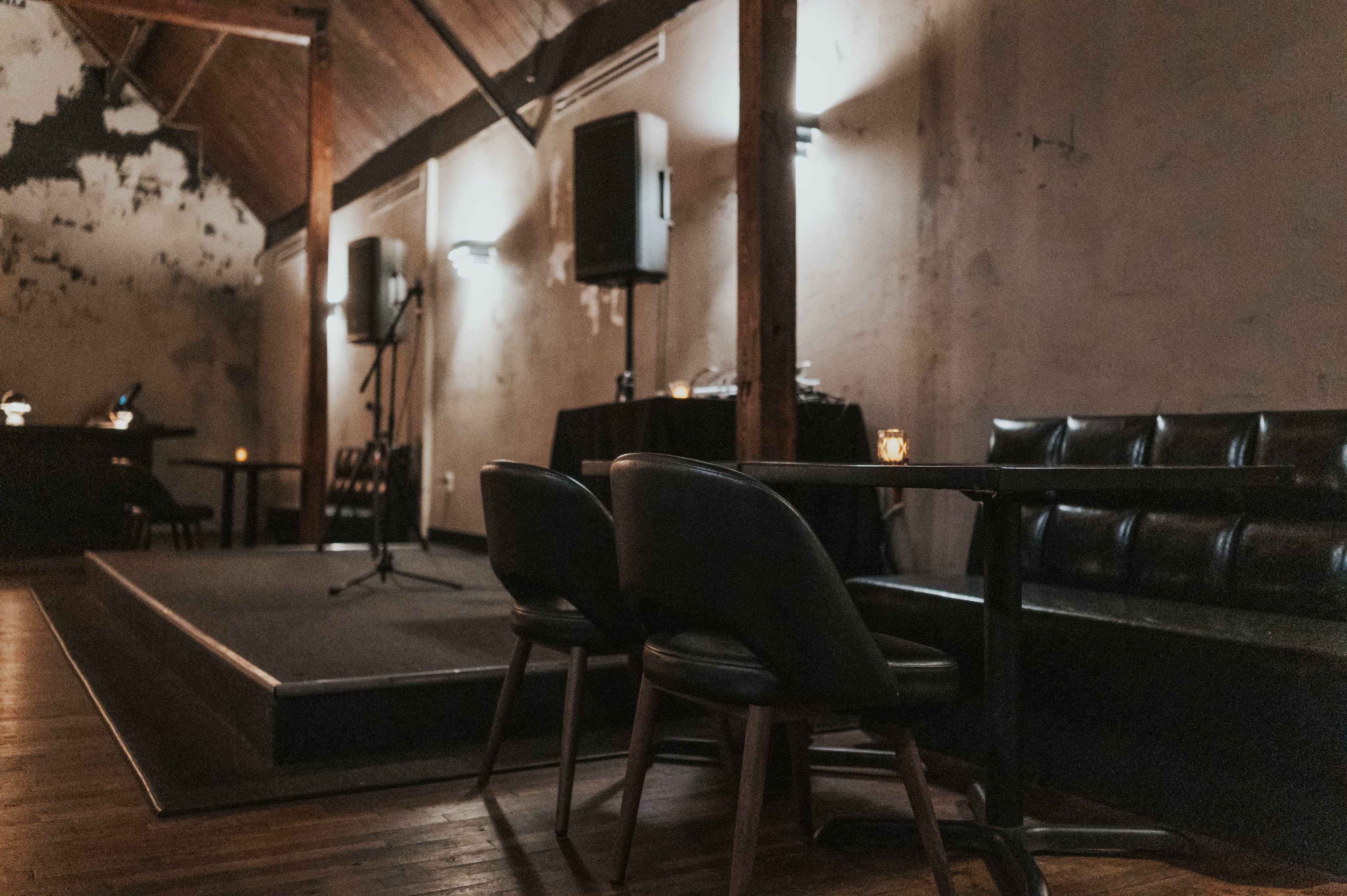 The image shows a dimly lit room with a small stage, a microphone stand, and seating arranged around dark wood tables.