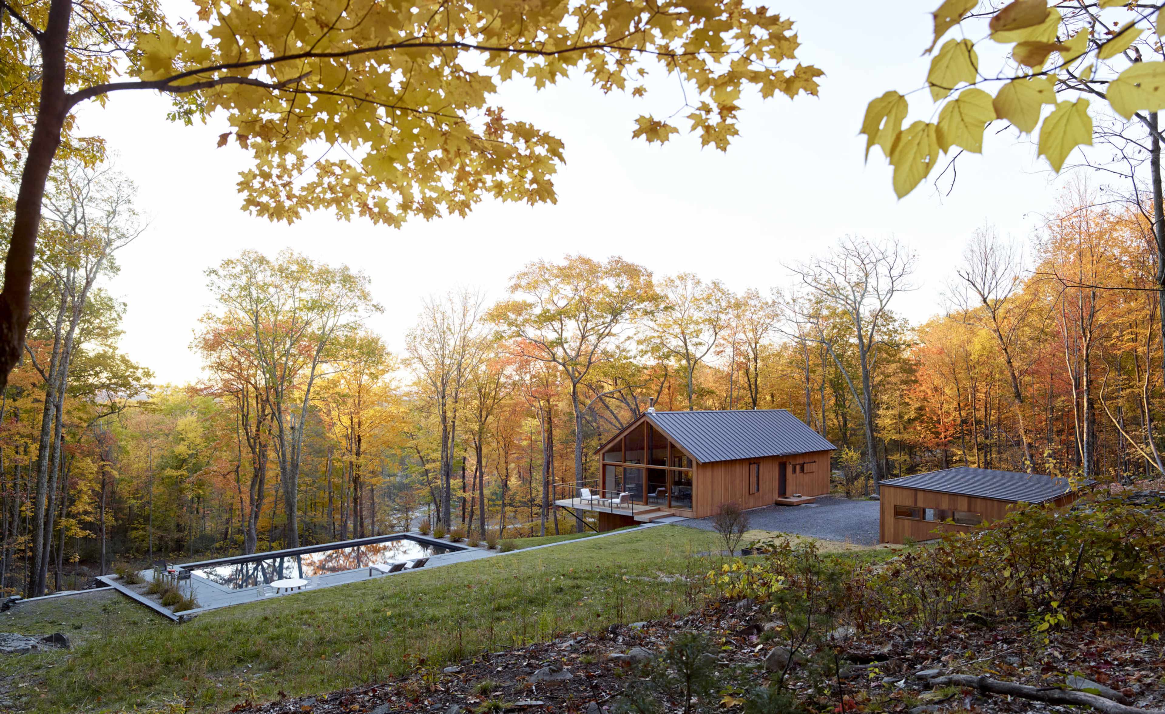 A wooden house with a gabled roof is situated near a rectangular pool, surrounded by trees with autumn foliage.