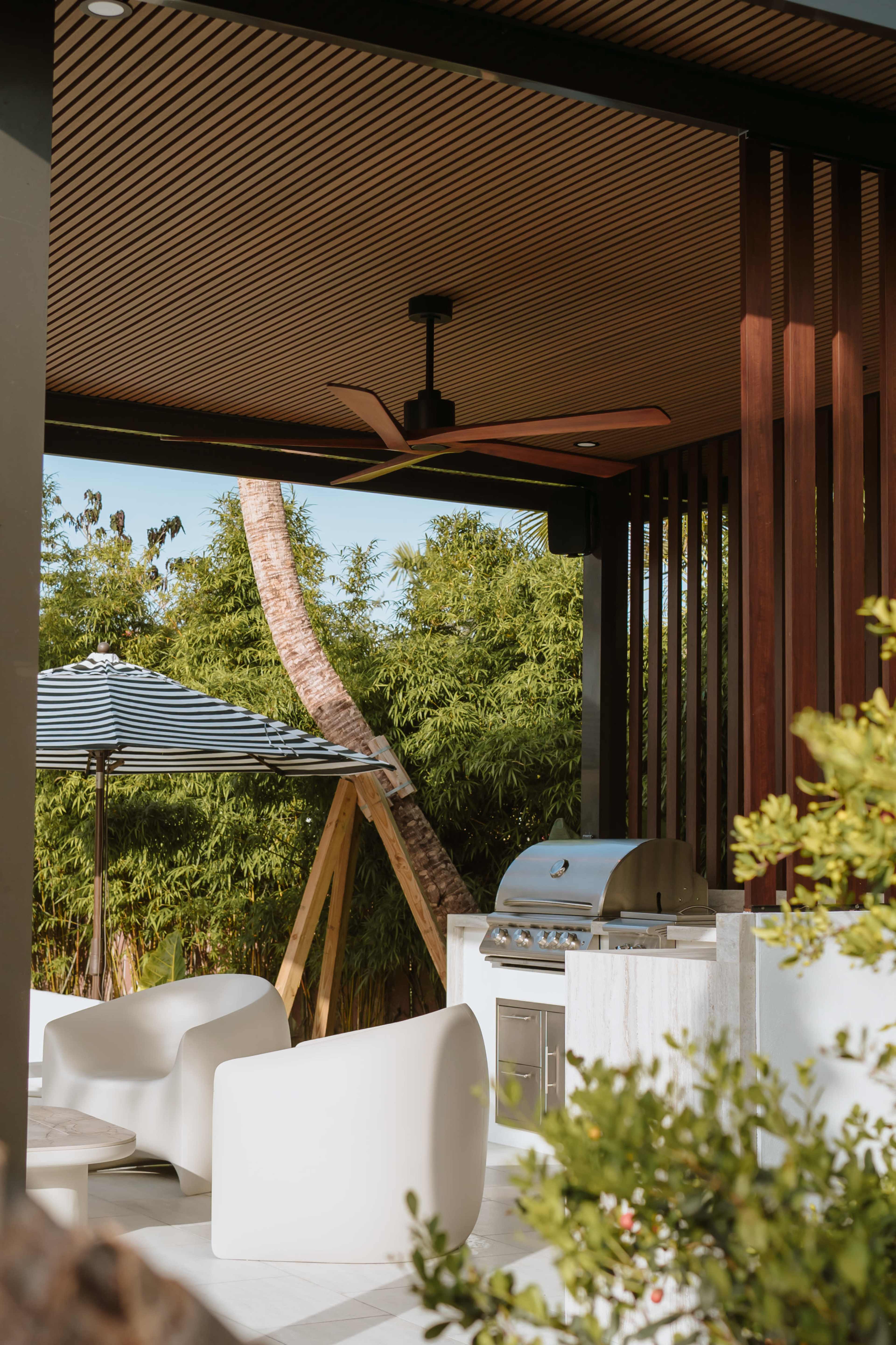 The image shows an outdoor grilling area featuring a stainless steel barbecue, modern white seating, and a fan under a wooden canopy surrounded by greenery.
