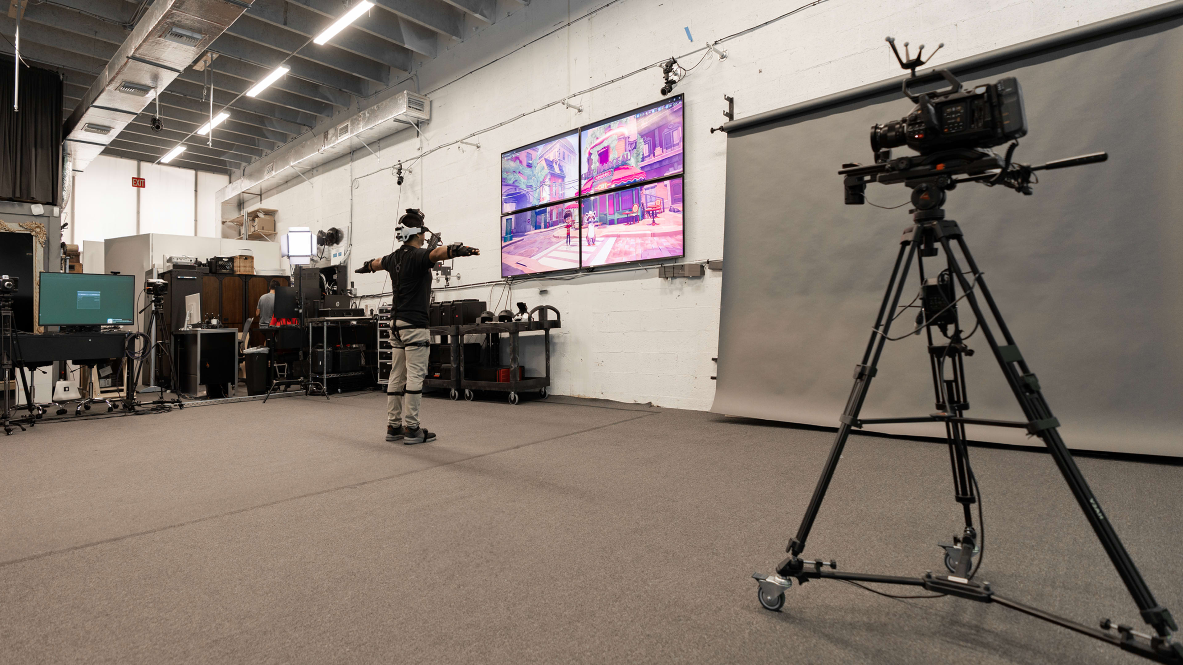 A person wearing a virtual reality headset stands in front of a large screen displaying a digital scene, while a camera on a tripod is positioned nearby.