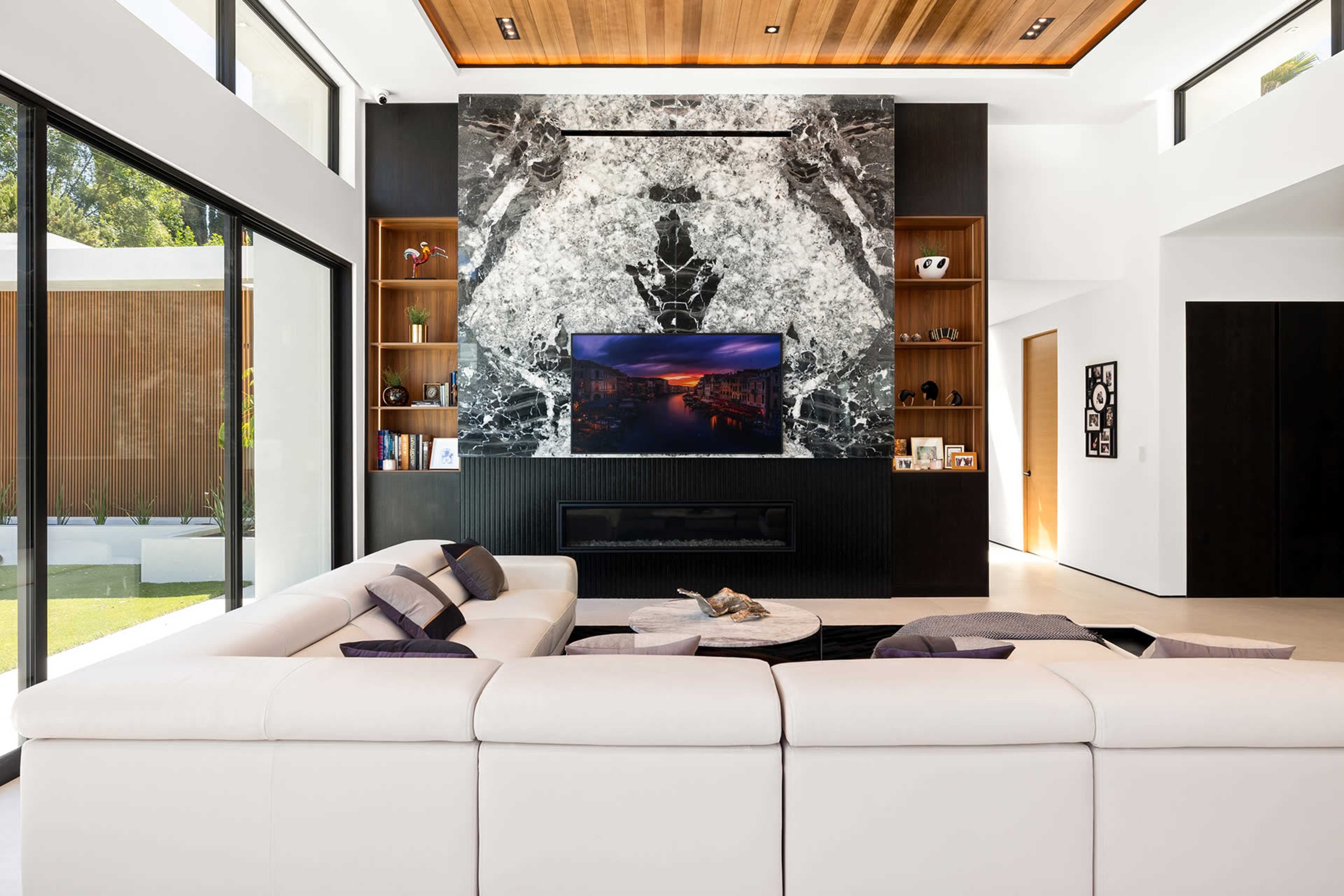 The image shows a modern living room with a large white sectional sofa, a decorative stone wall featuring a television, and large windows providing natural light.