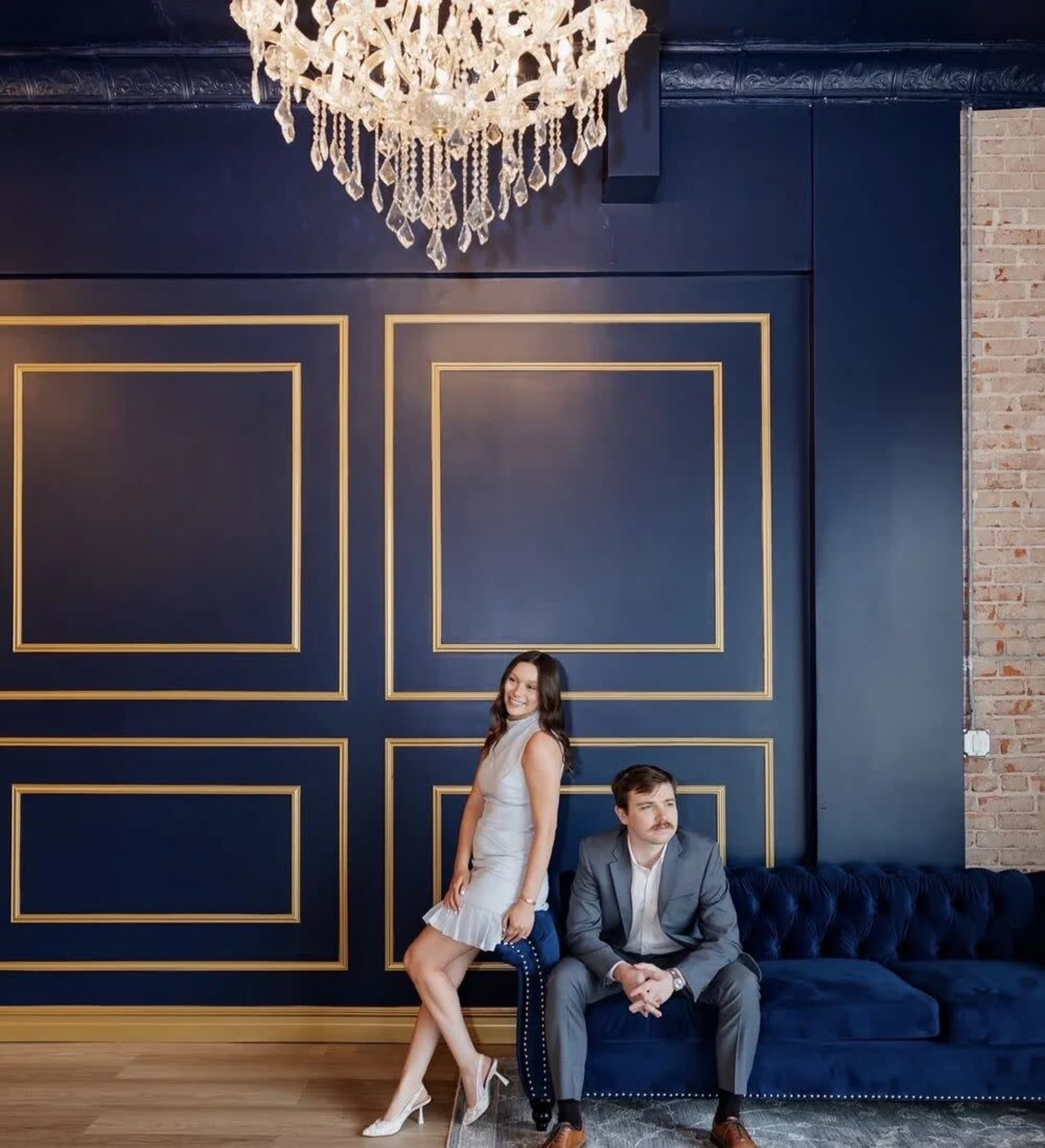A man in a suit and a woman in a light dress pose in front of a dark blue wall with gold accents and a chandelier overhead.
