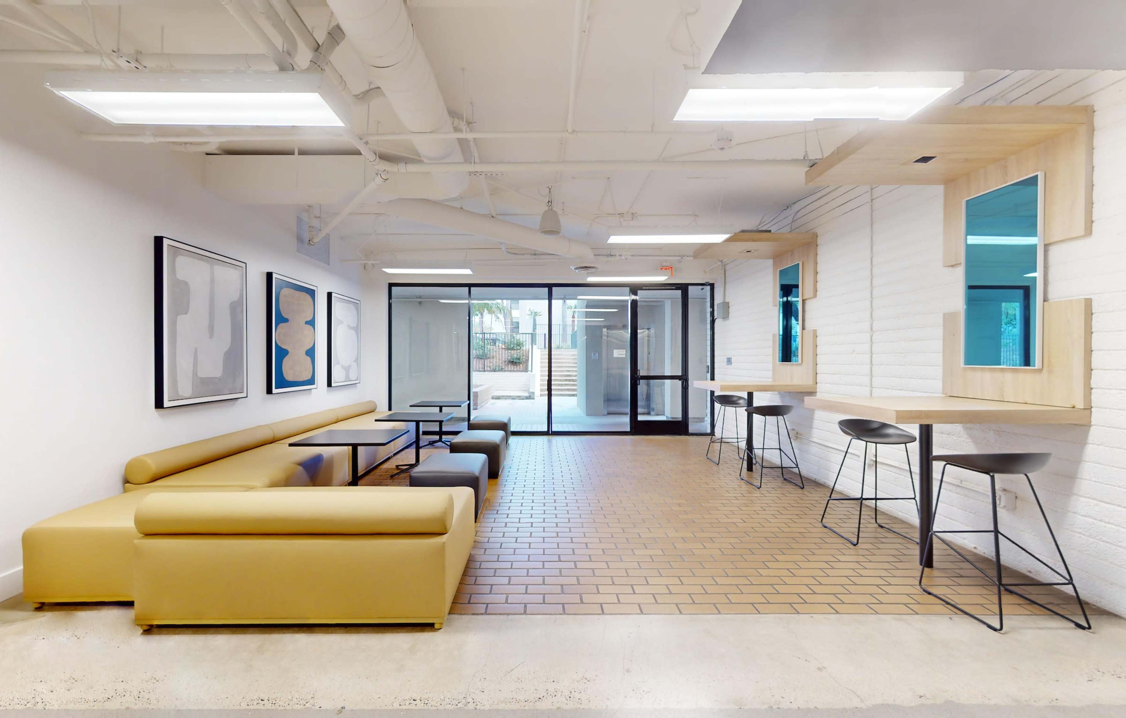 A modern indoor seating area featuring a yellow sofa, black tables, and tall stools against a backdrop of large glass doors leading to an outdoor space.