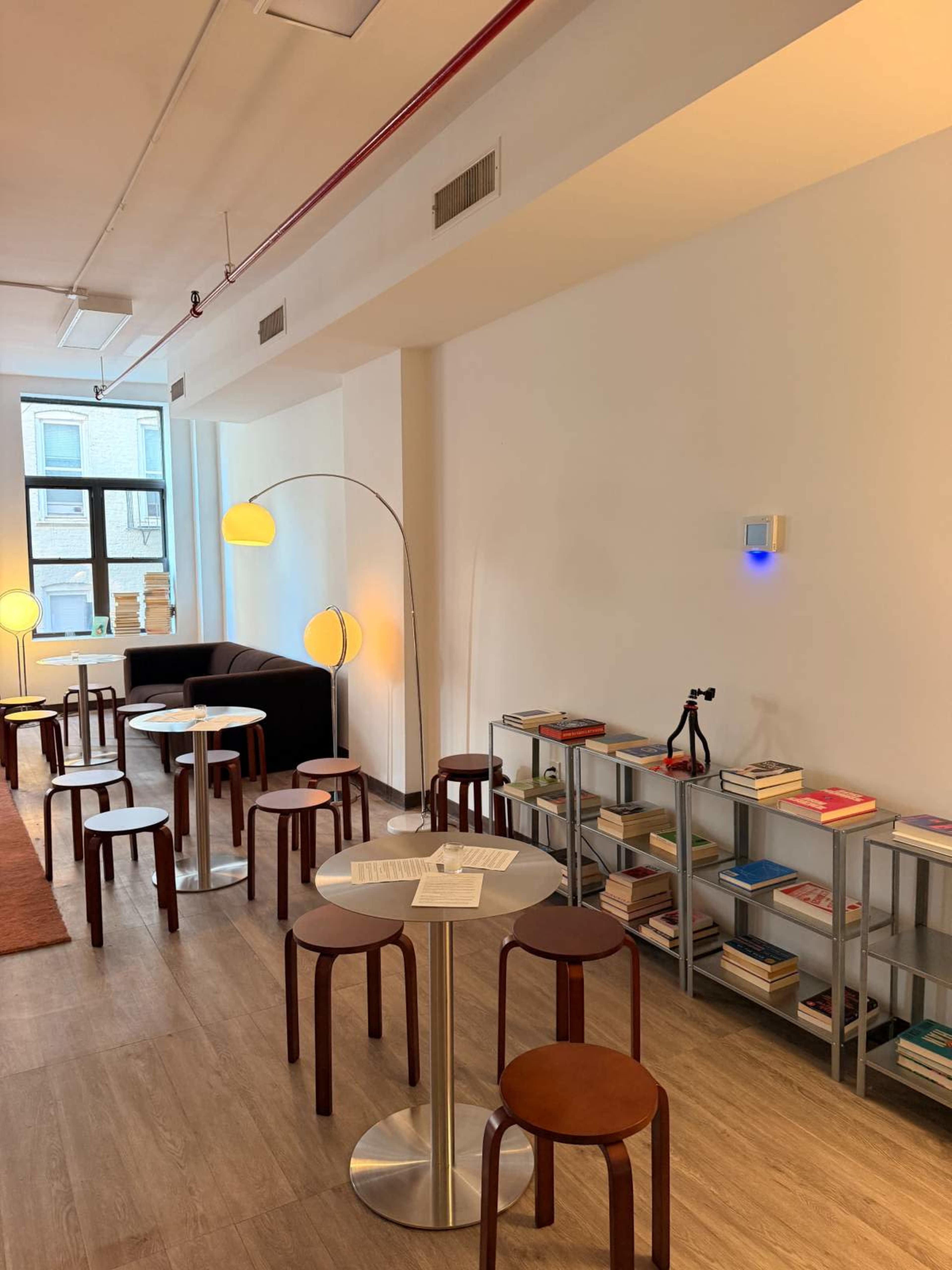 The image shows a modern, minimalist café space with circular tables, wooden stools, a sofa, and shelves lined with books.