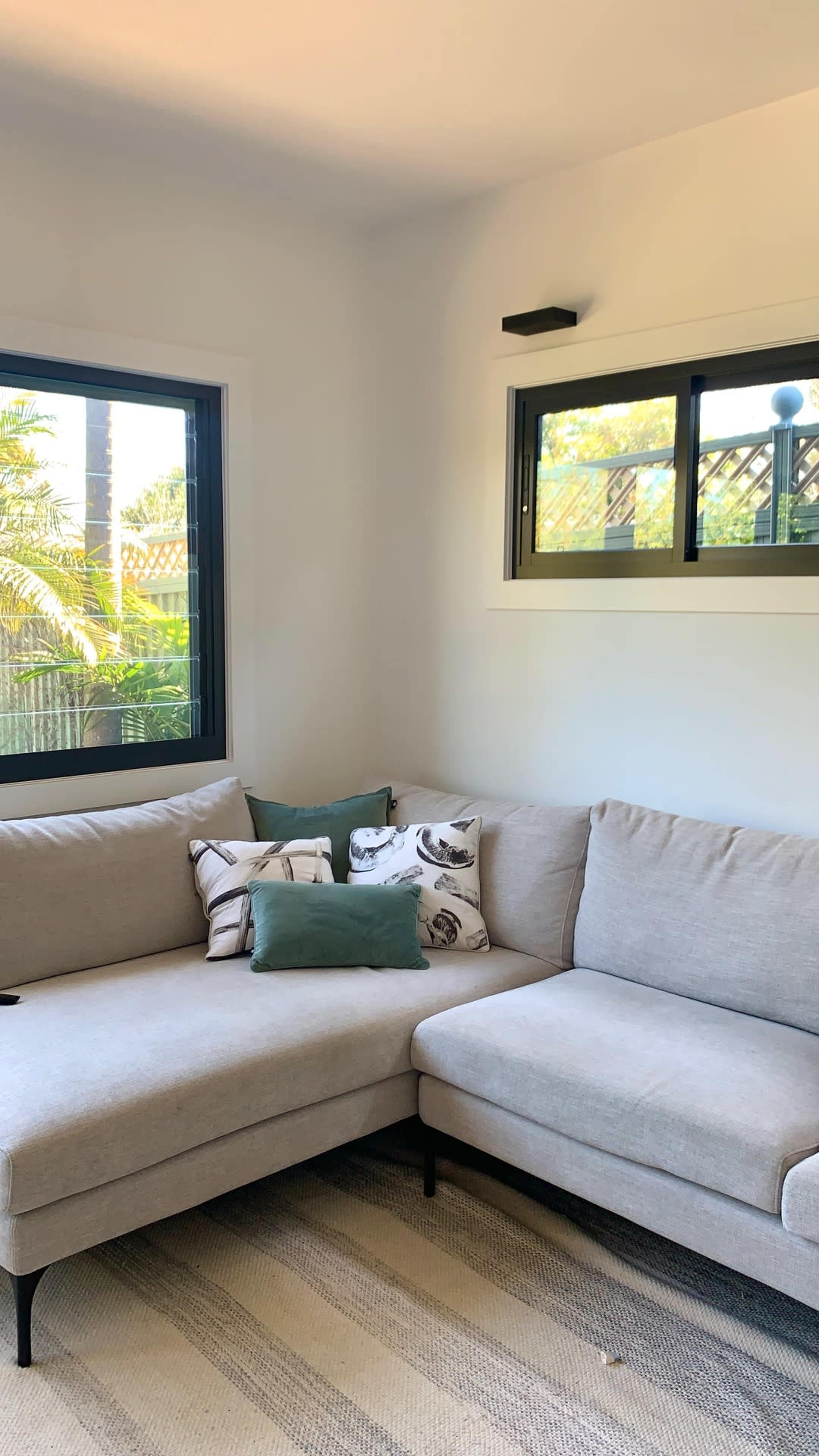 The image shows a light-colored sectional sofa in a bright room with large windows and decorative pillows on the cushions.