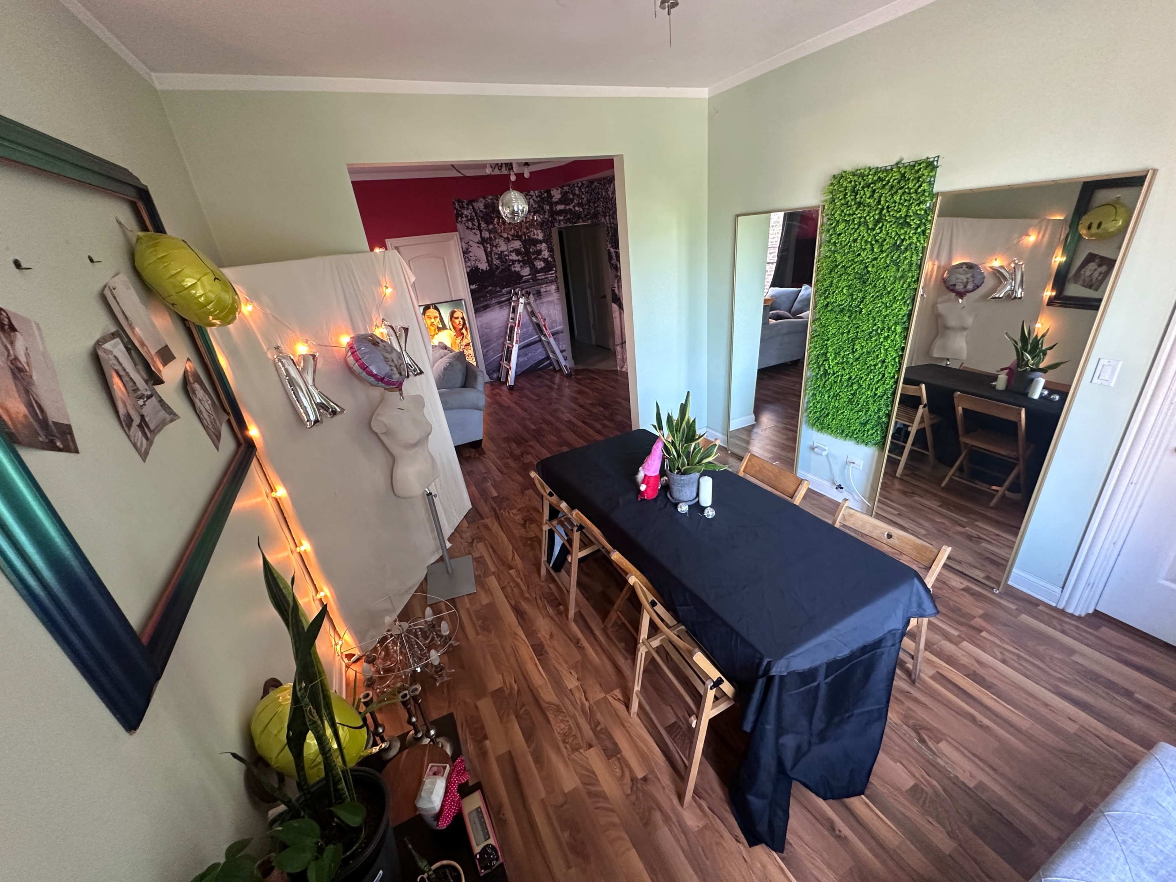 A dining area features a long black table surrounded by wooden chairs, with decorative plants and lights in a brightly lit room.