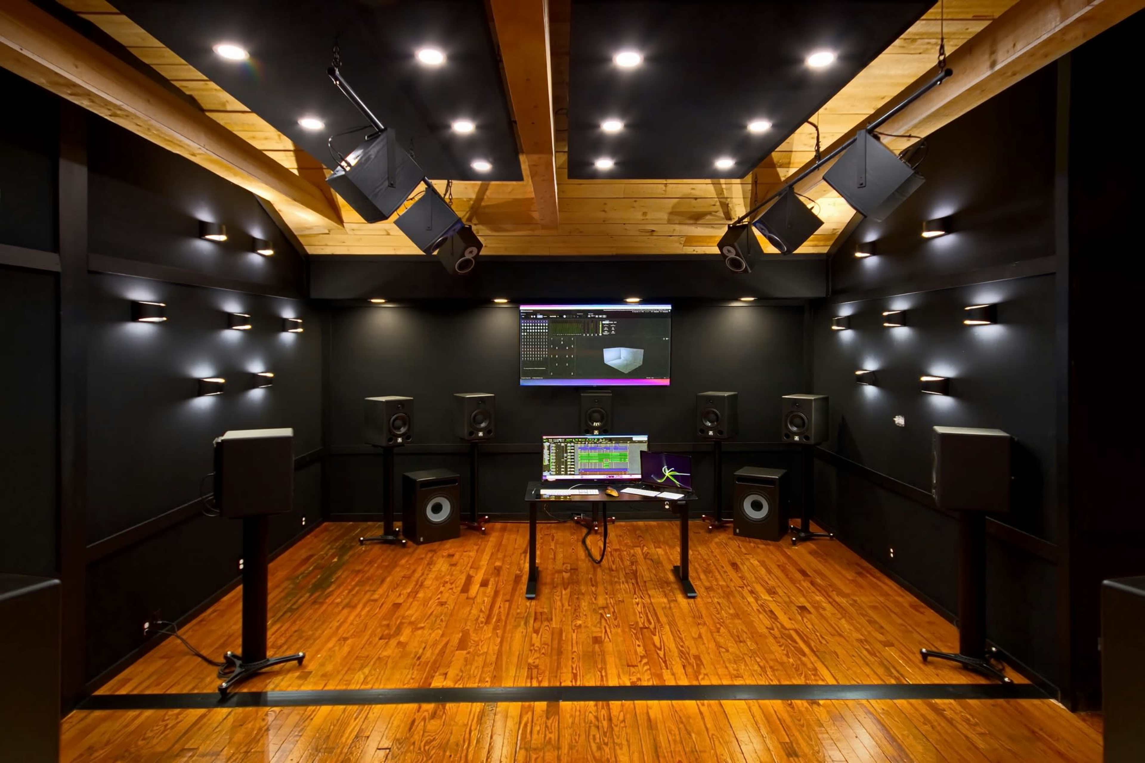 A soundproof audio studio with a wooden floor, equipped with speakers, a mixing table, and a large monitor displaying audio software.