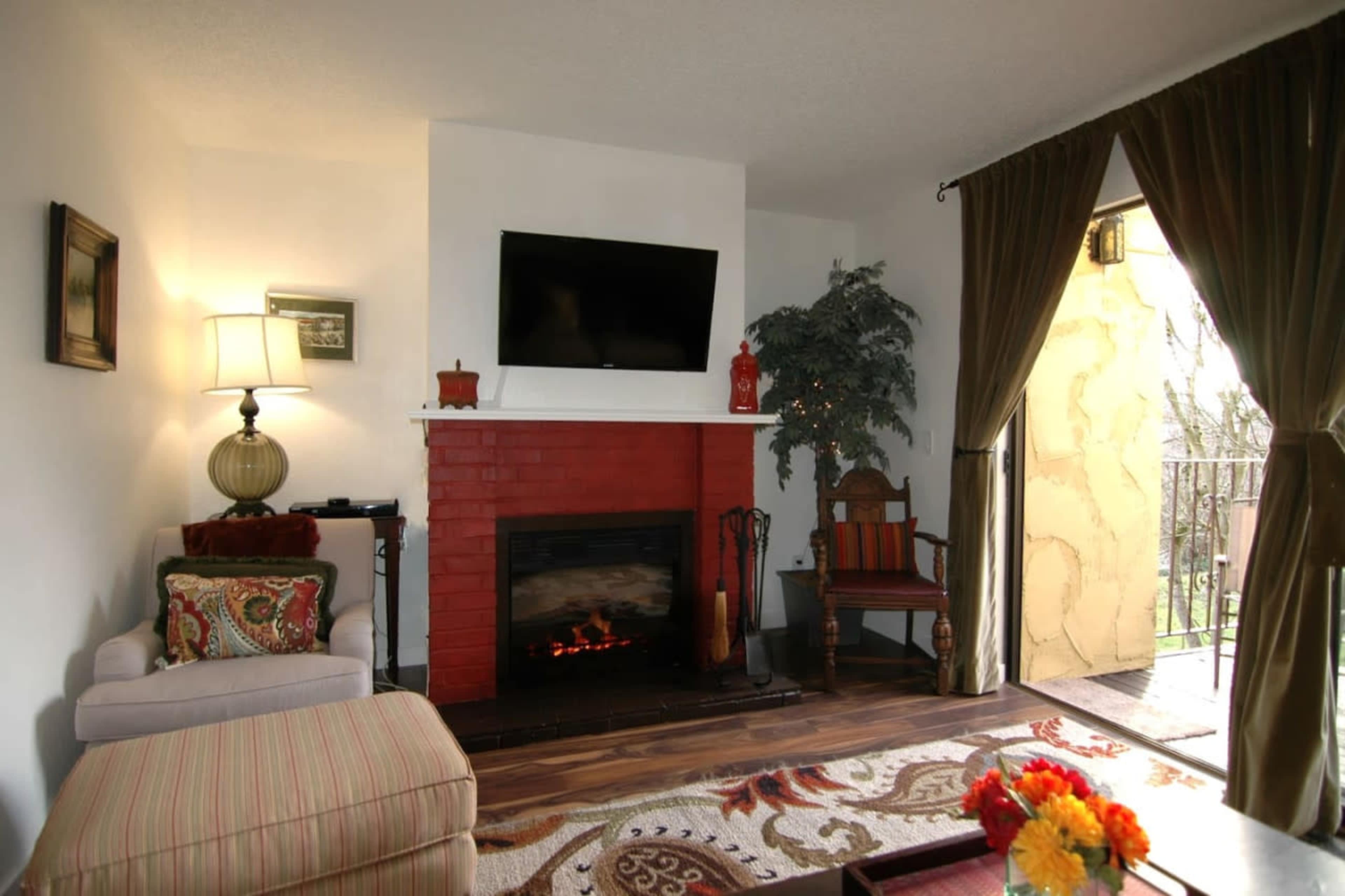 The image shows a neatly arranged living room featuring a fireplace with a TV above it, a comfortable chair, and decorative plants.