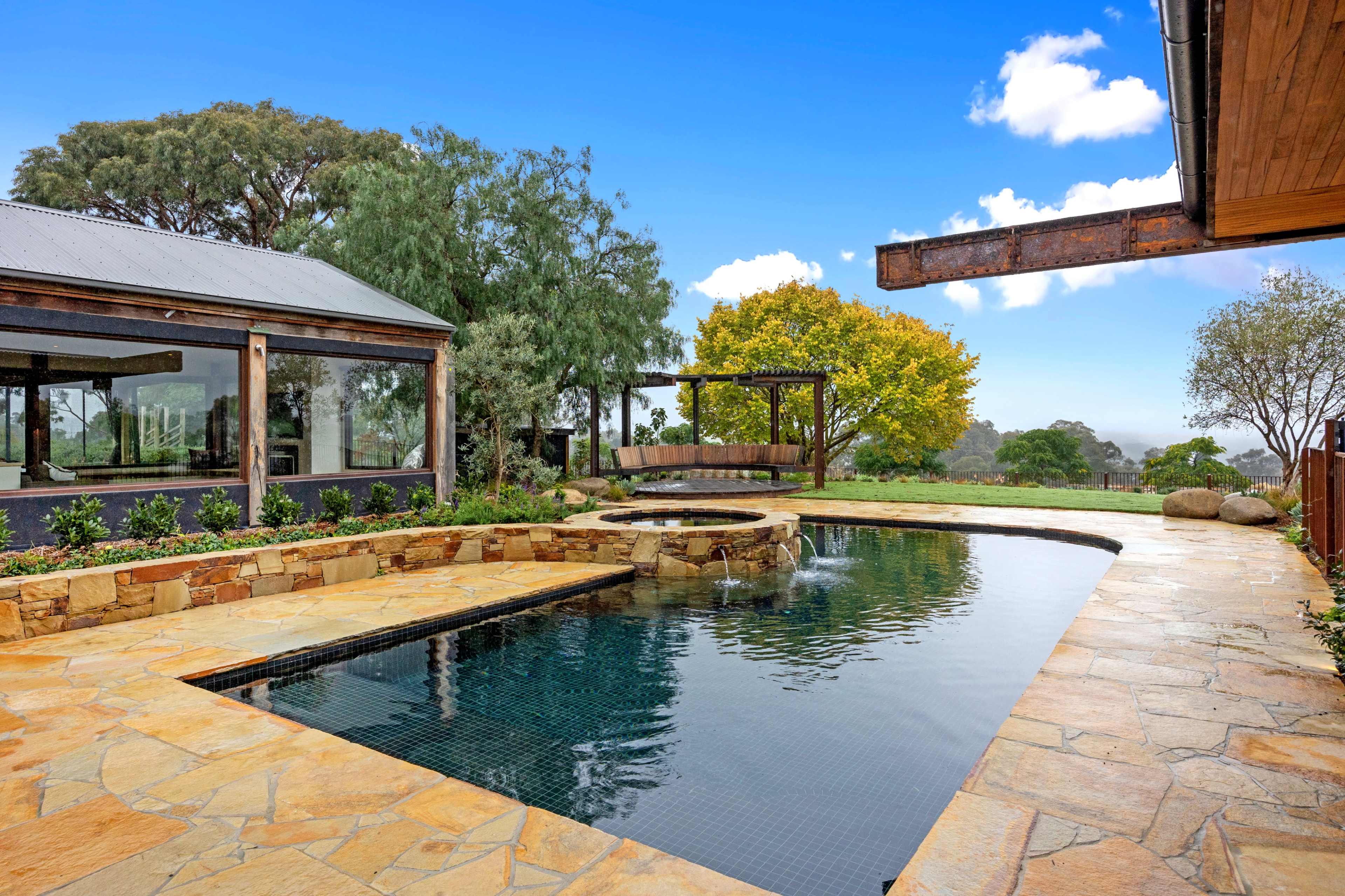 A stone patio with a swimming pool and a swing set surrounded by trees and a clear sky.