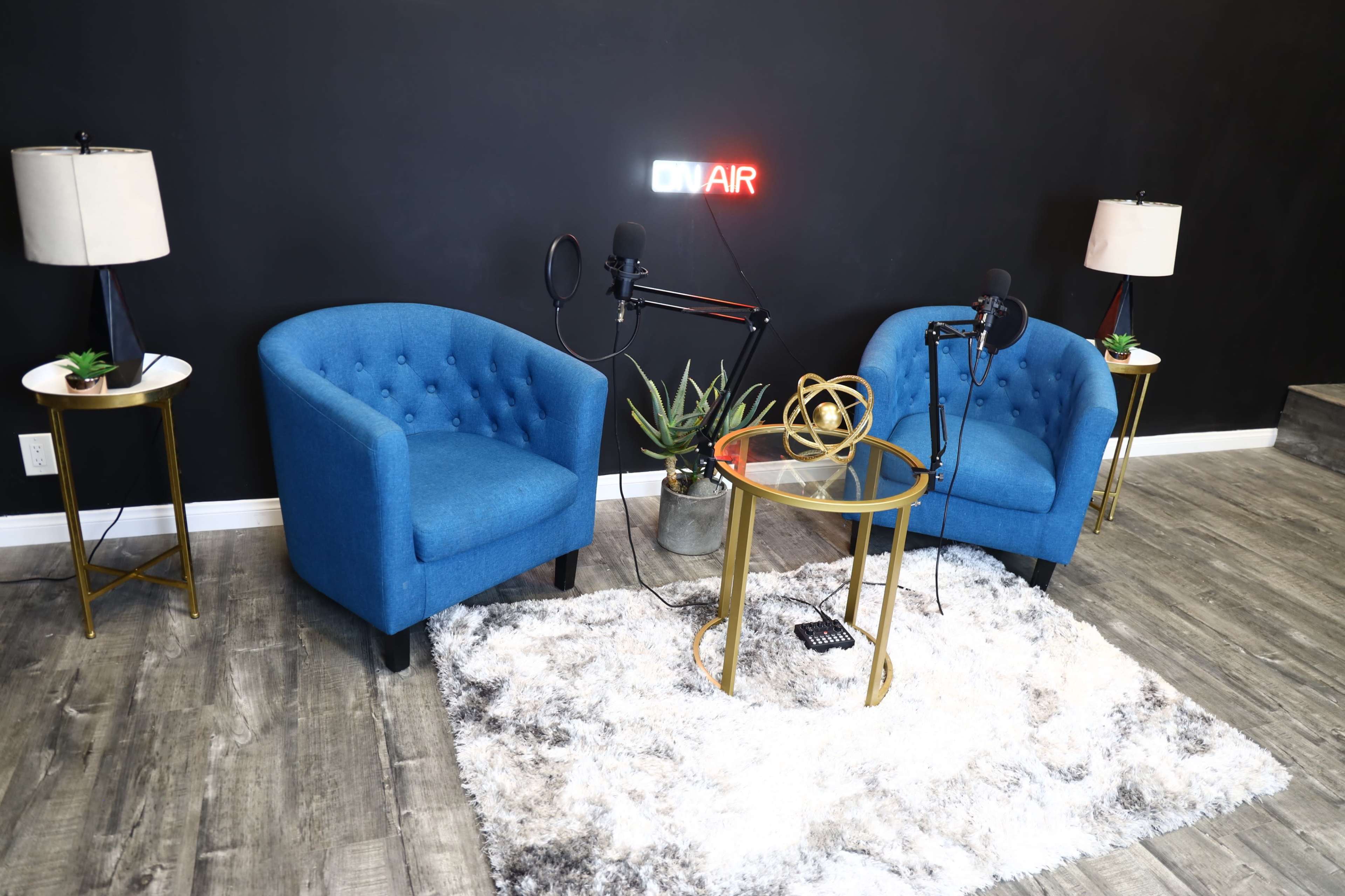 The image shows a podcast setup featuring two blue chairs, a glass table with decorative items, and microphones on stands, all against a black wall with an "ON AIR" sign.