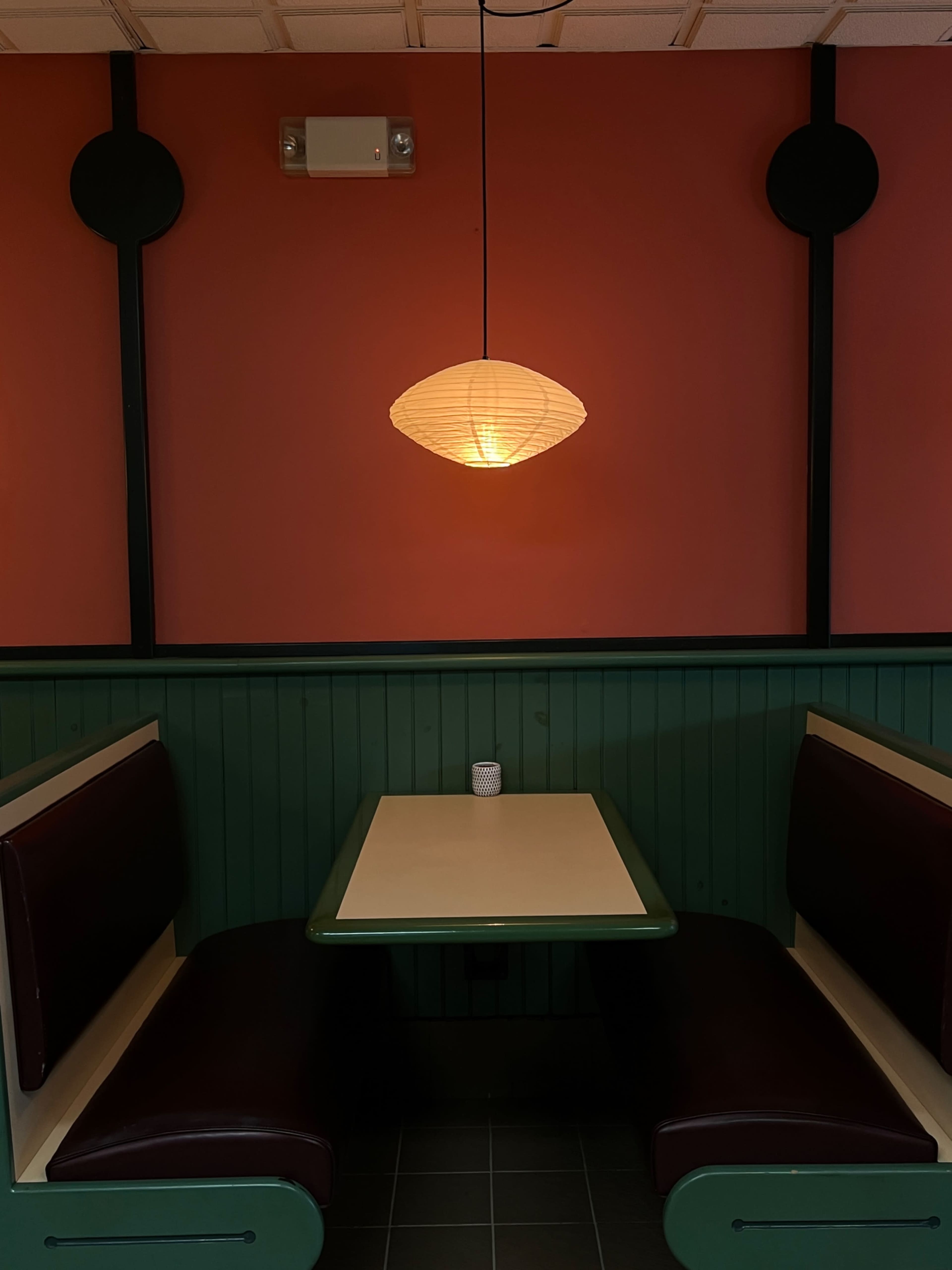 A cozy booth with a green and white table, dark brown seating, and a pendant light hanging above, set against a red wall.