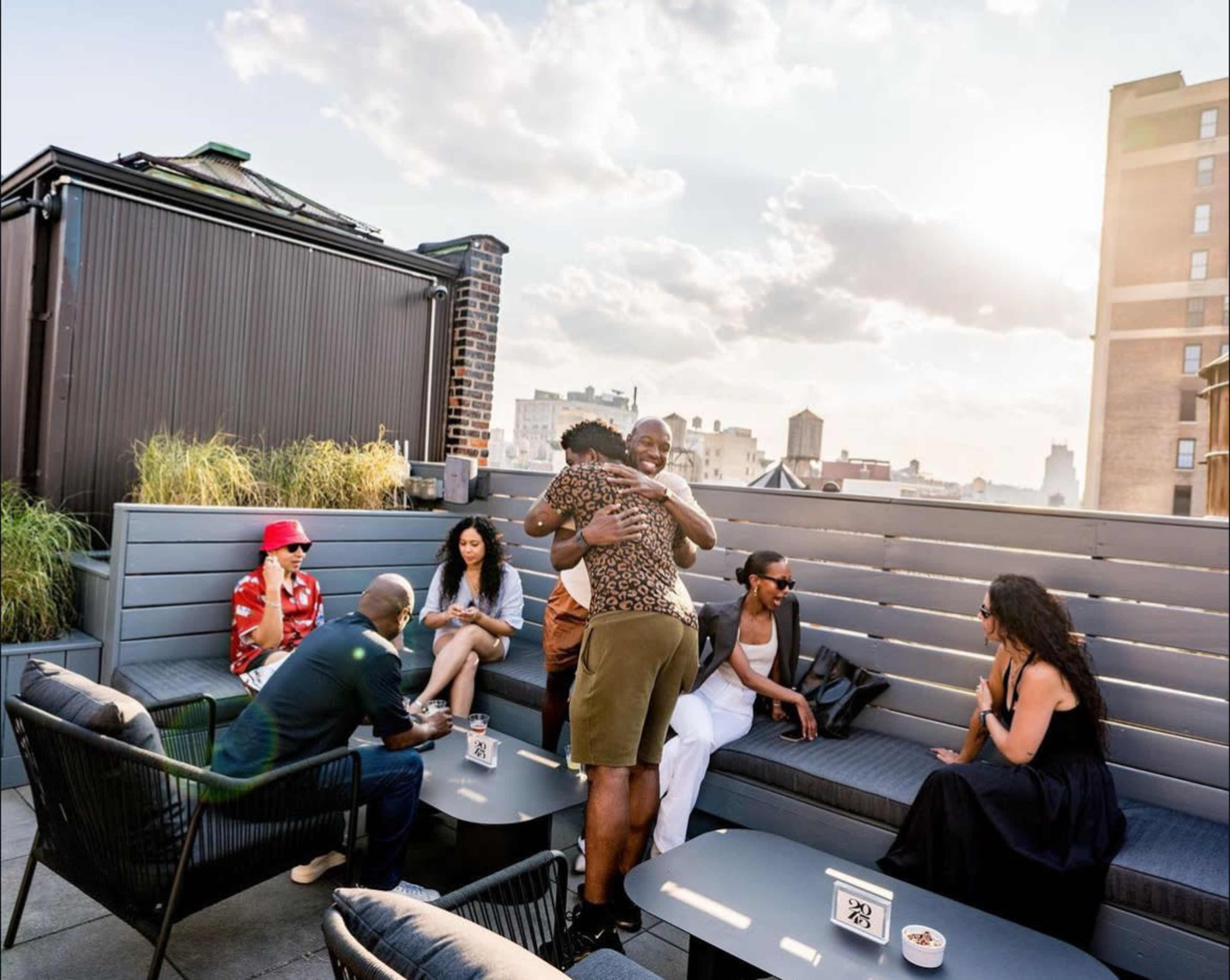 A group of people socializes on a rooftop, some seated at tables while others embrace, with a skyline and clouds visible in the background.