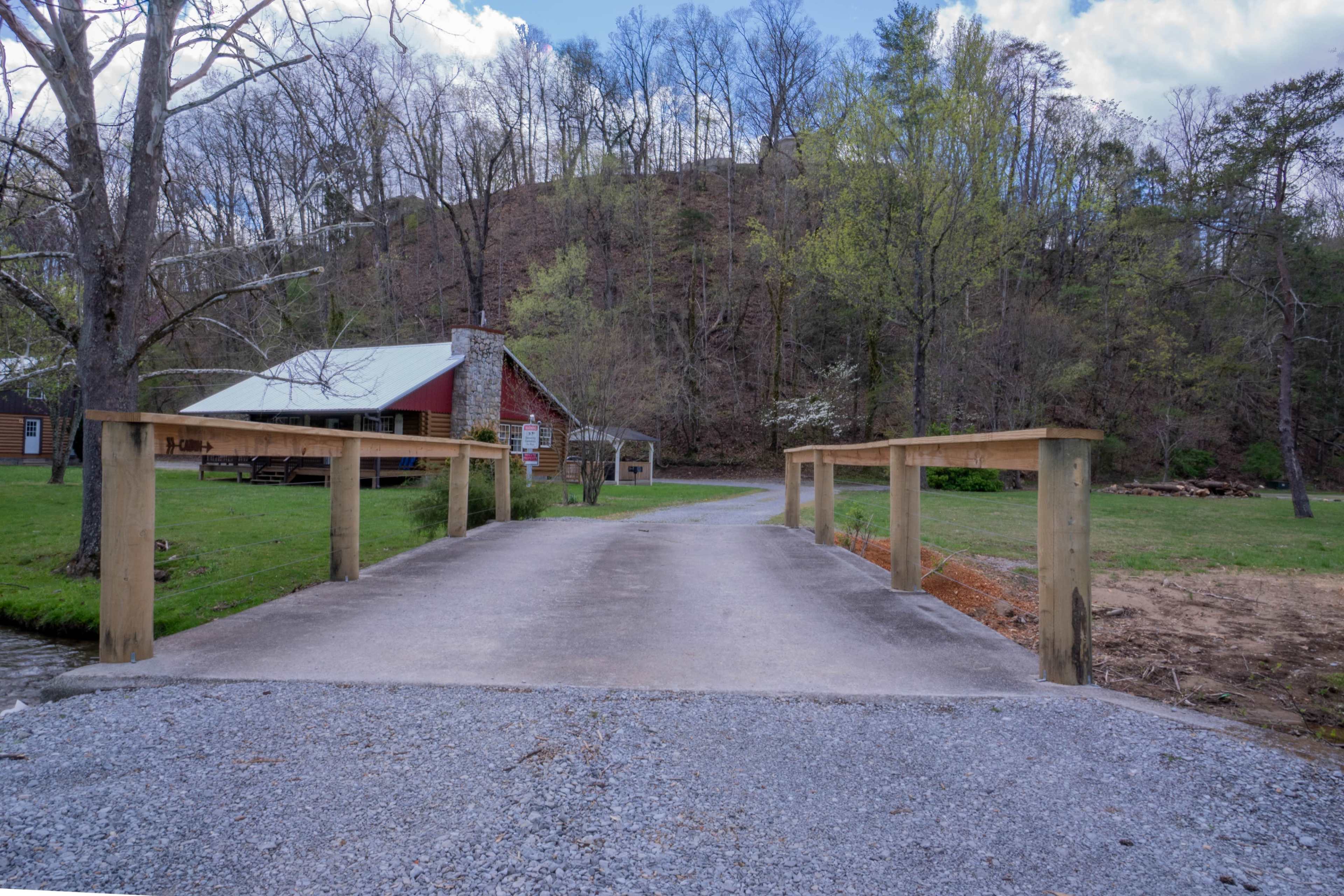 A concrete bridge leads to a cabin situated on a grassy area, surrounded by trees and a hillside in the background.