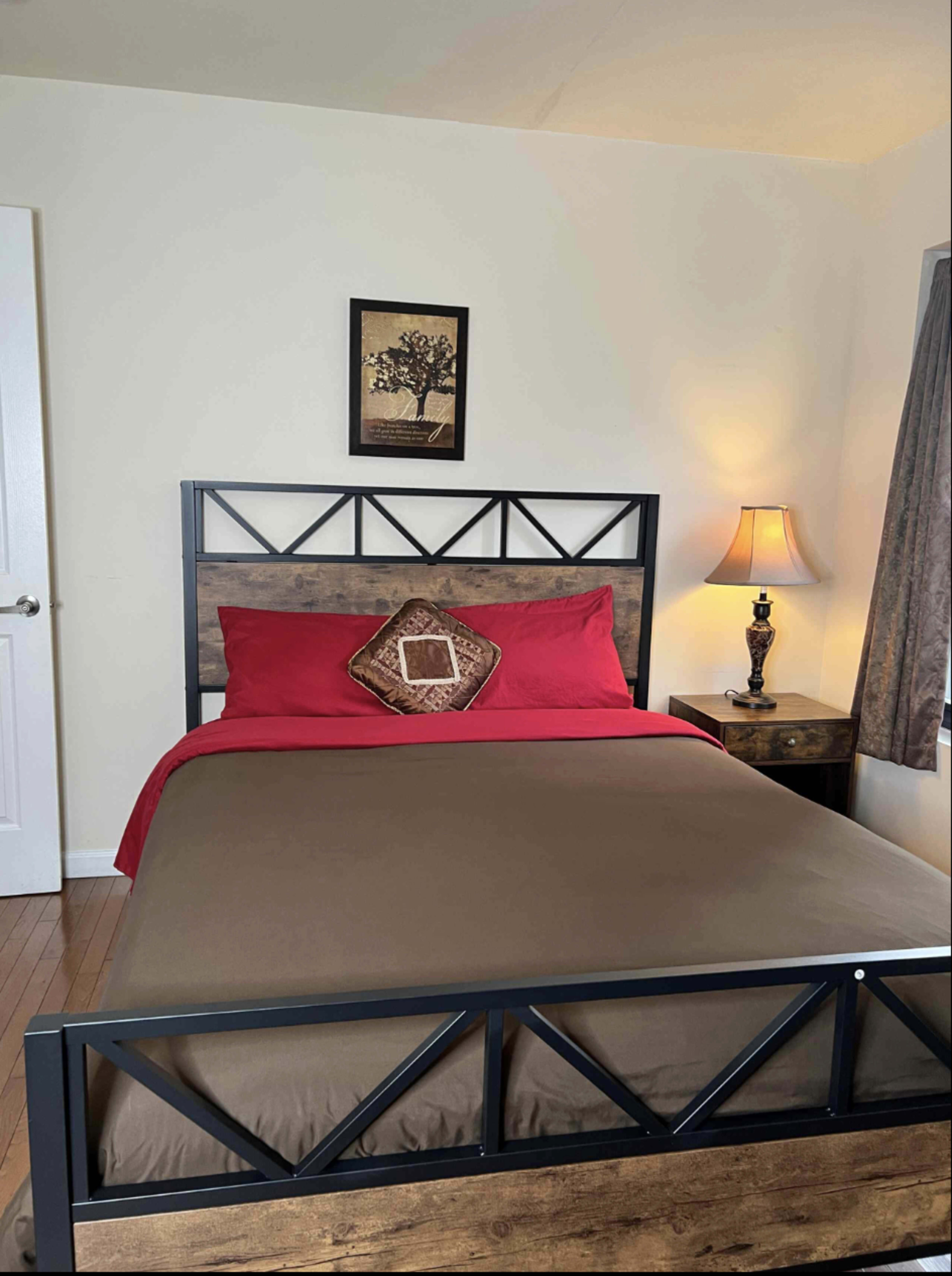 The image shows a neatly arranged bedroom featuring a bed with a red and brown color scheme, a bedside table with a lamp, and a framed artwork on the wall.