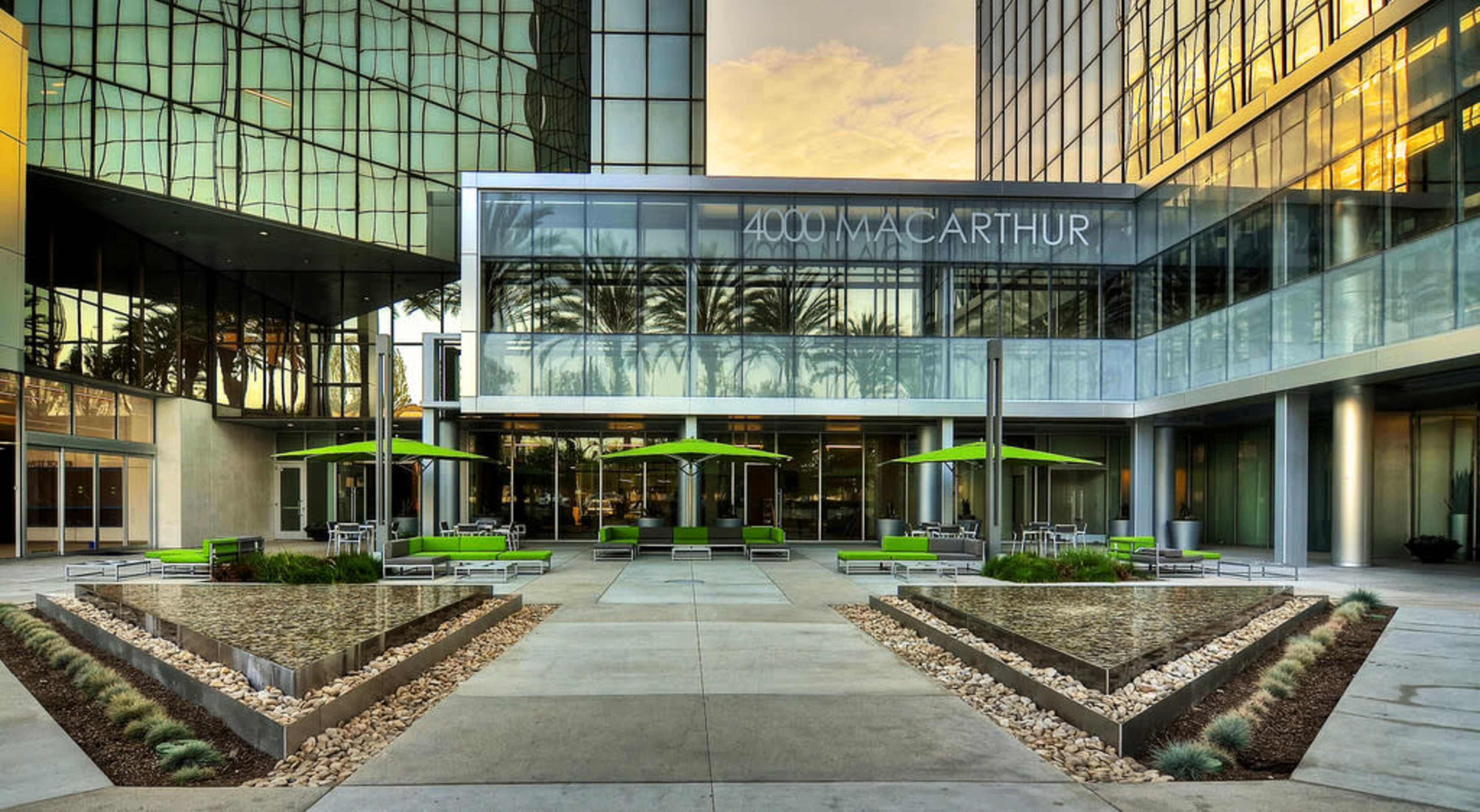 The image depicts a modern office building entrance with a landscaped plaza featuring green seating areas and umbrellas, framed by large glass structures.