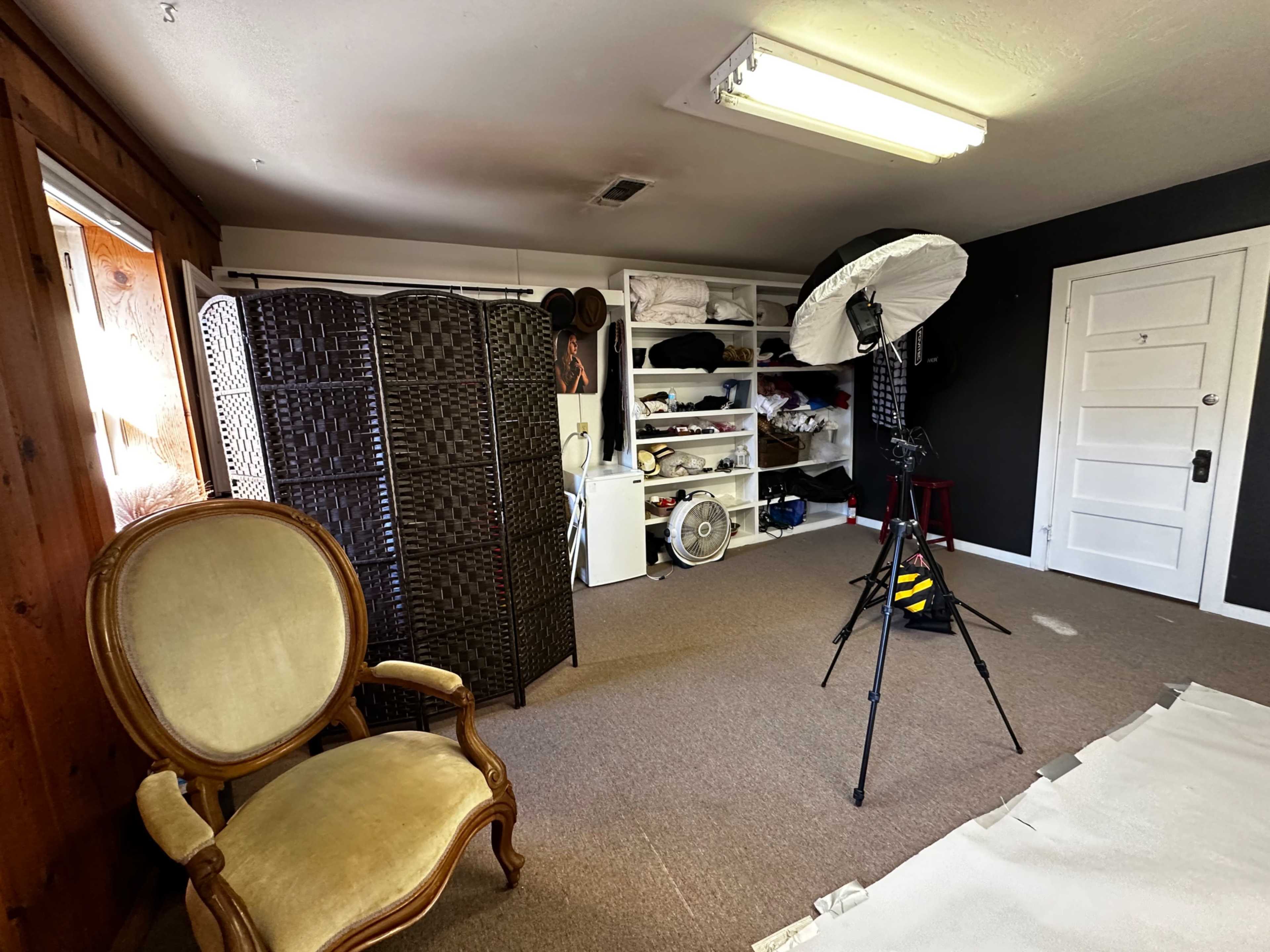 A well-lit room features a vintage chair, a folding room divider, shelves with various items, and photography equipment set up against a dark wall.