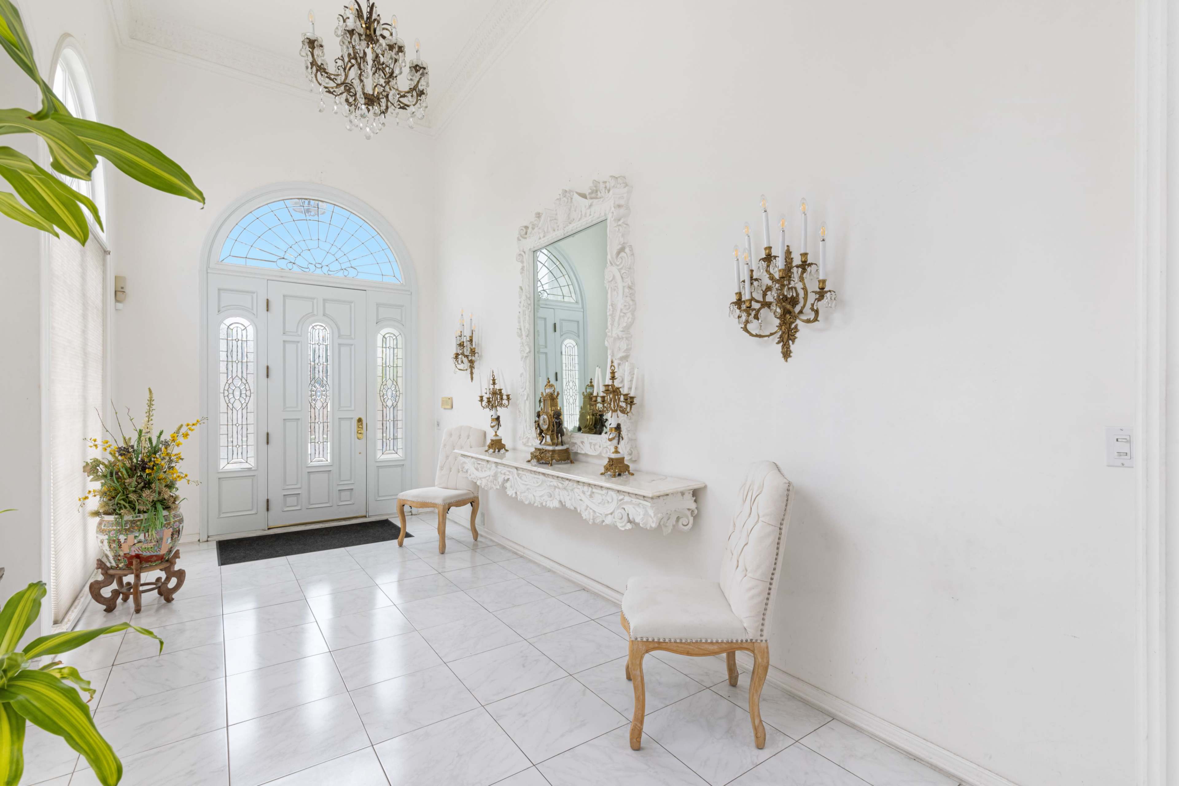 The image shows a bright, white foyer featuring a pair of double doors, a large mirror, wall sconces, and decorative chairs.
