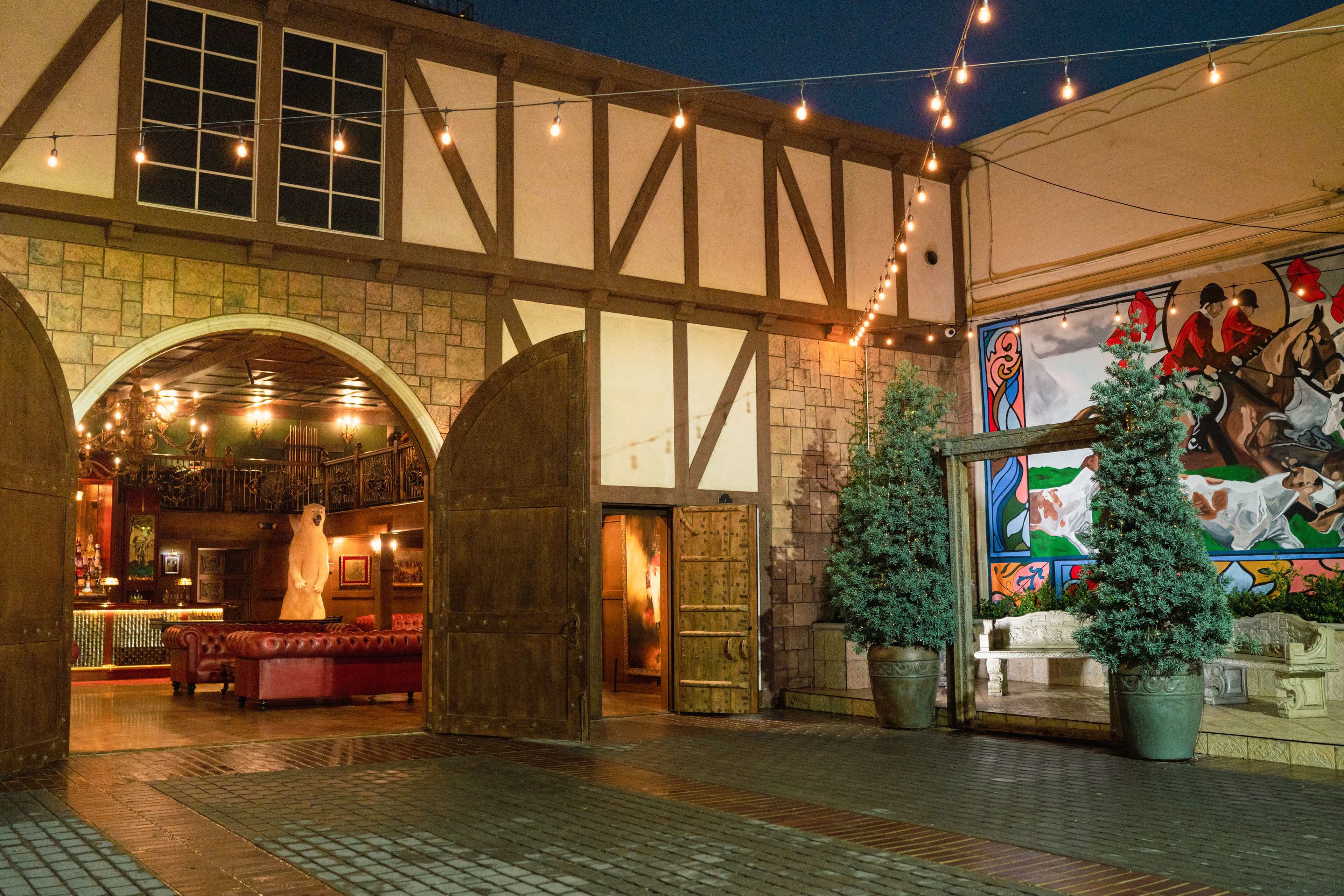 A courtyard area featuring wooden doors, a vintage red sofa, and decorated walls with artwork under twinkling string lights at dusk.