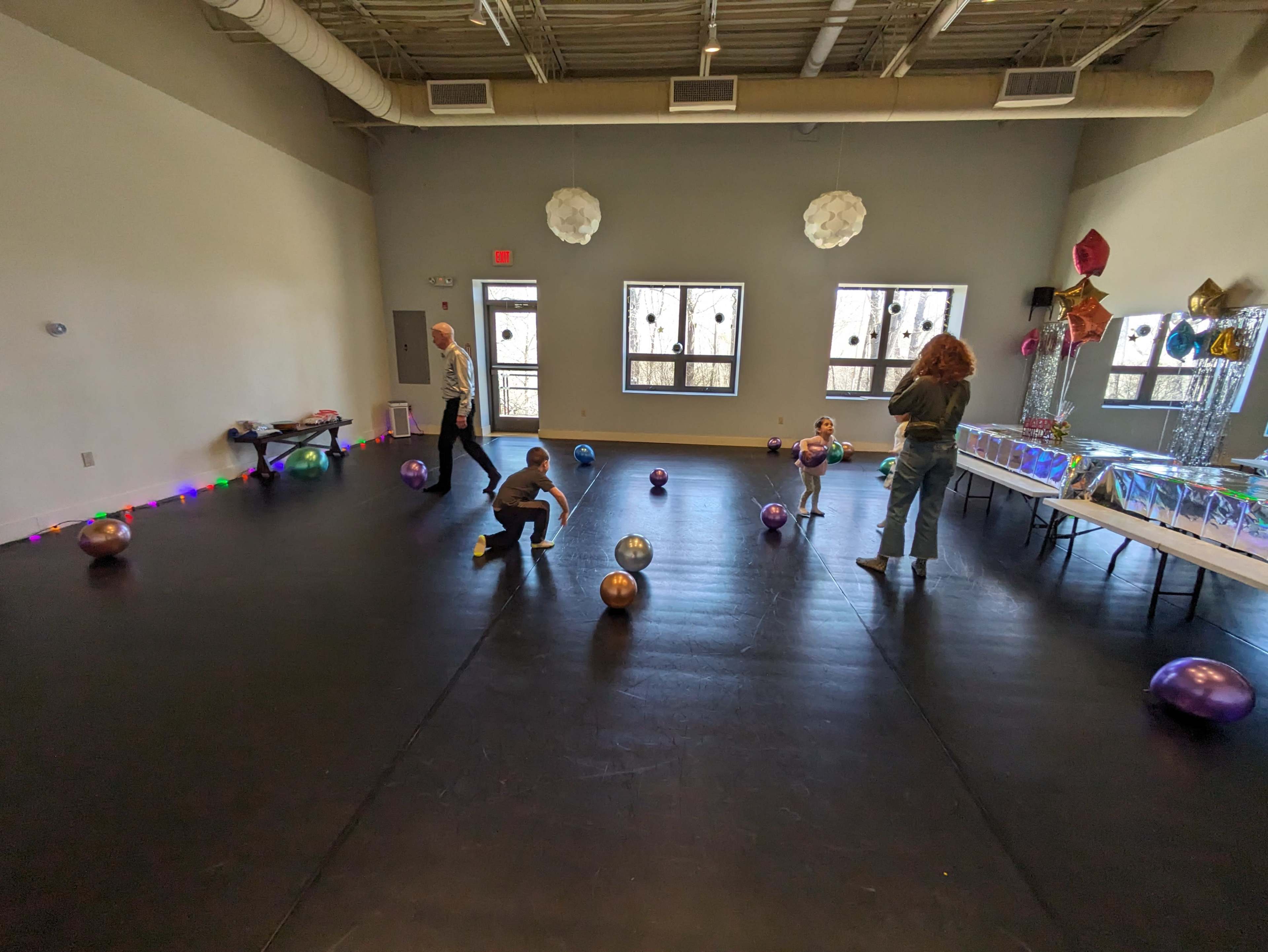 A spacious room with a black floor, two adults and three children are playing with colorful balloons near a table set up for an event, with decorations on the walls.