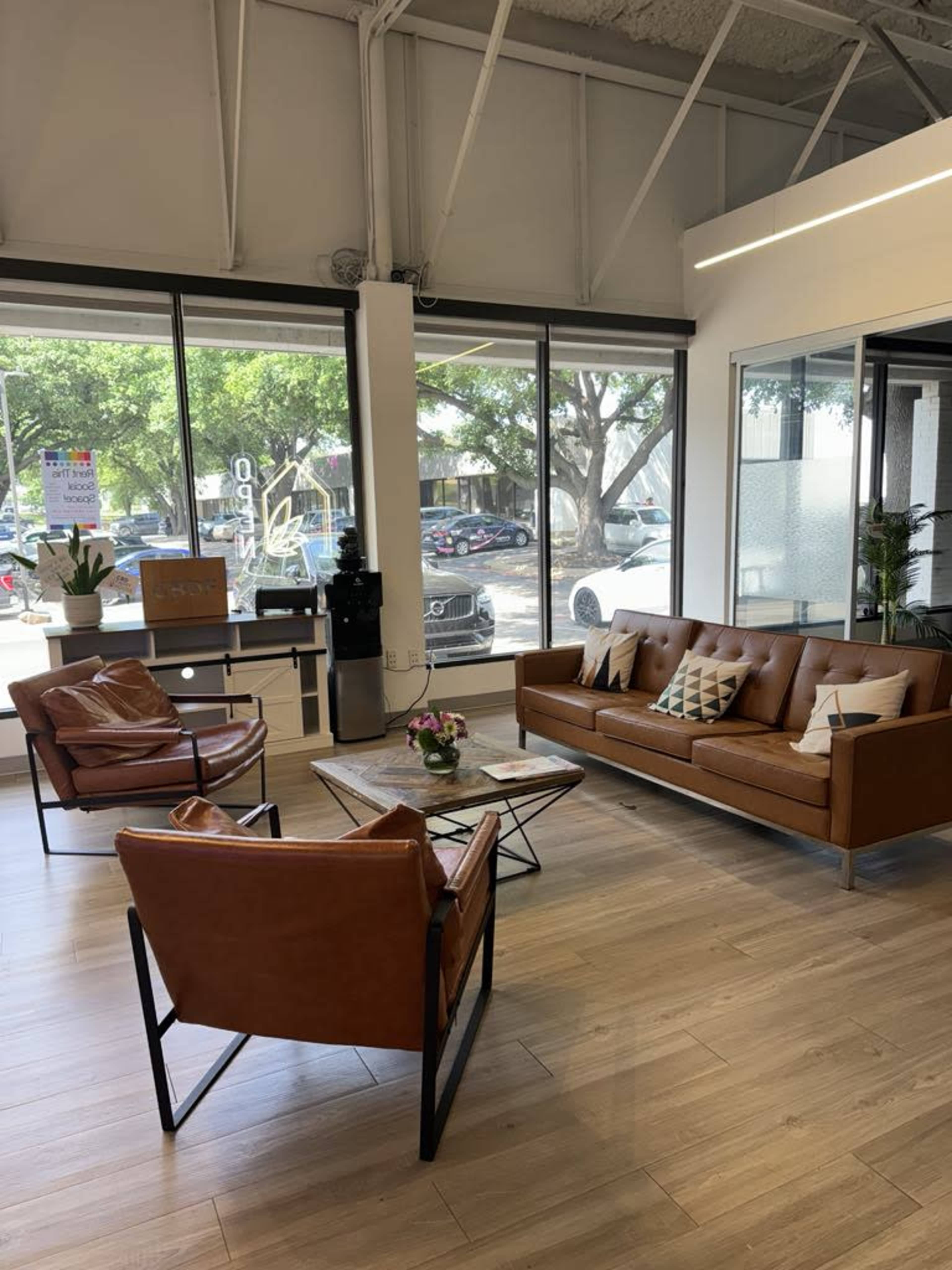 A modern office interior with brown leather furniture, including two chairs and a sofa, arranged around a coffee table, in front of large windows overlooking cars parked outside.