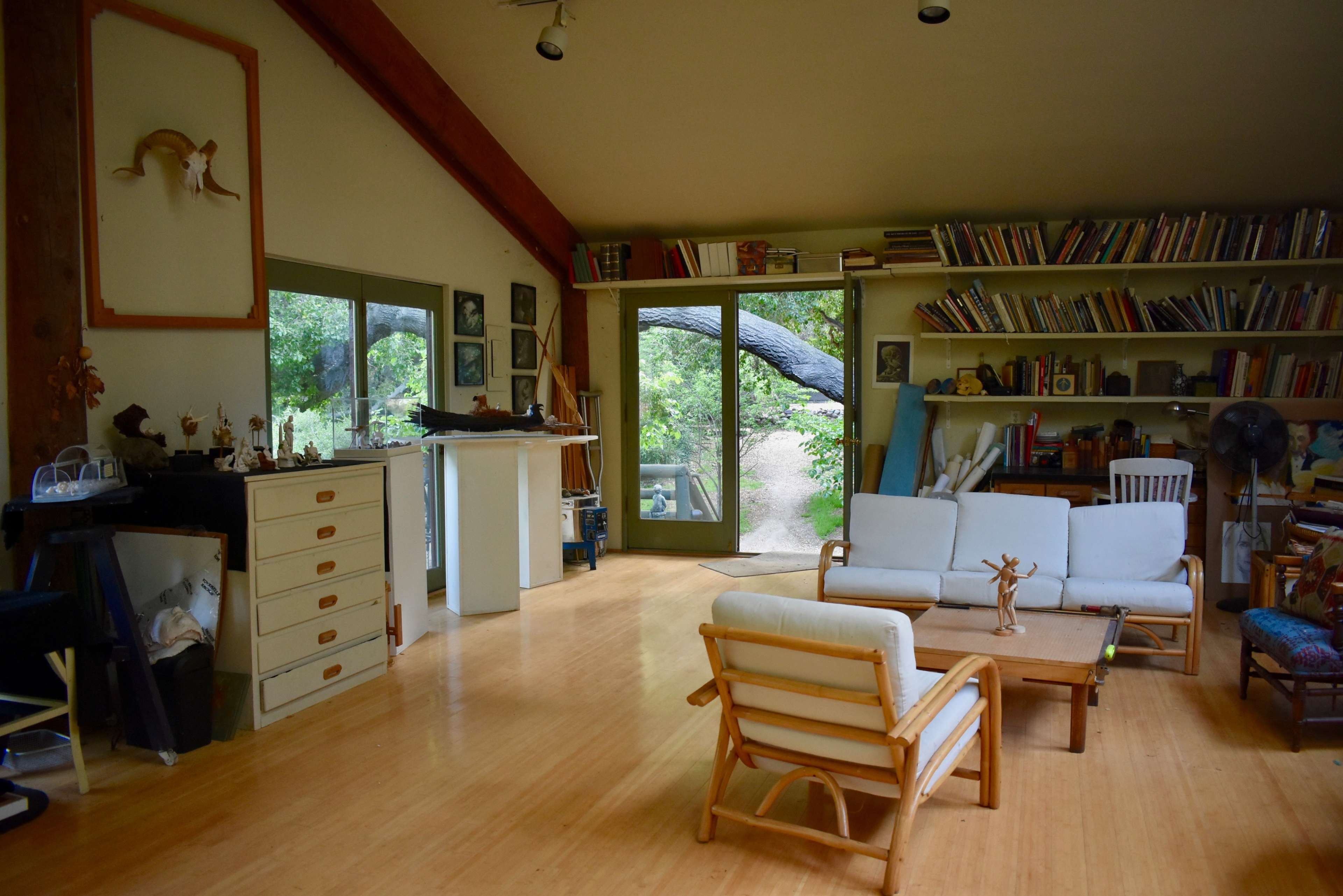 A spacious, well-lit living room with wooden floors, a large window opening to an outdoor area, and bookshelves filled with books and decorative items.