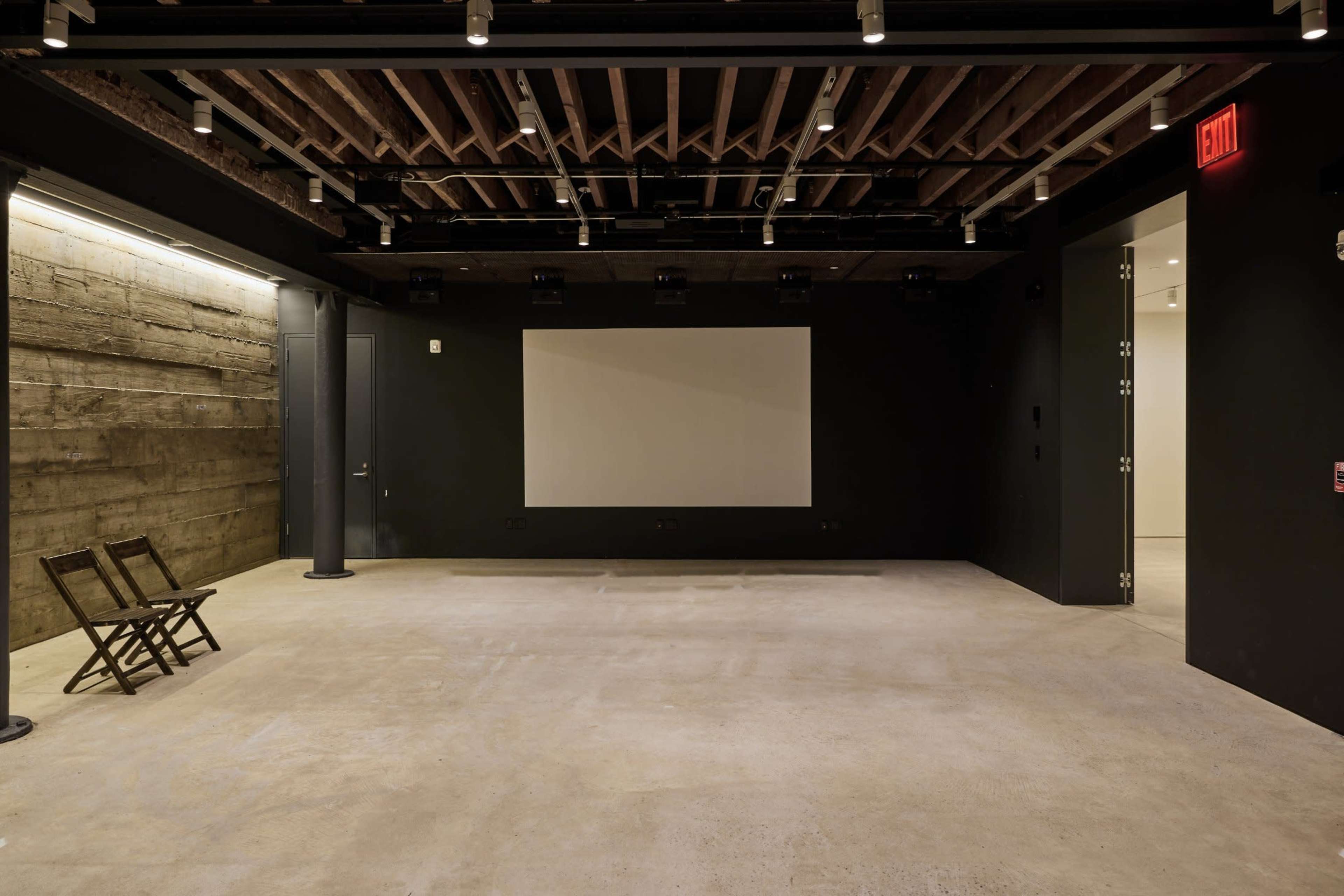 The image shows a minimalist, empty room with a concrete floor, exposed wooden beams, and a large blank screen on a black wall.