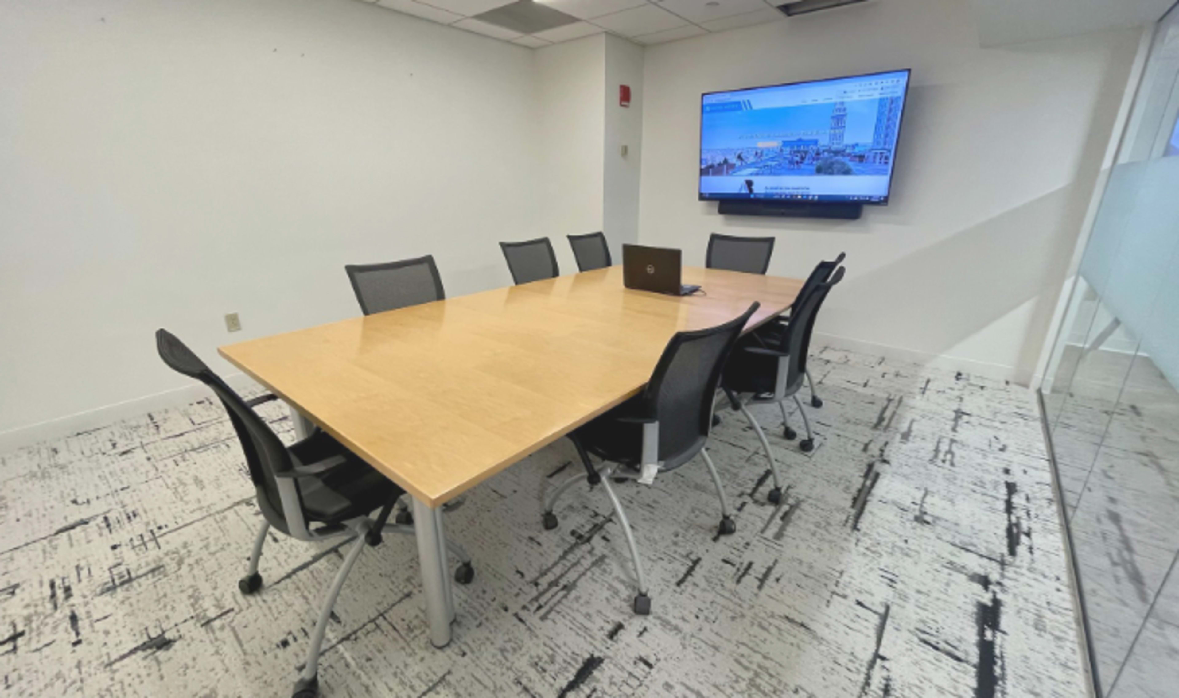The image shows a conference room featuring a large wooden meeting table surrounded by eight black mesh chairs, with a wall-mounted screen displaying a cityscape.