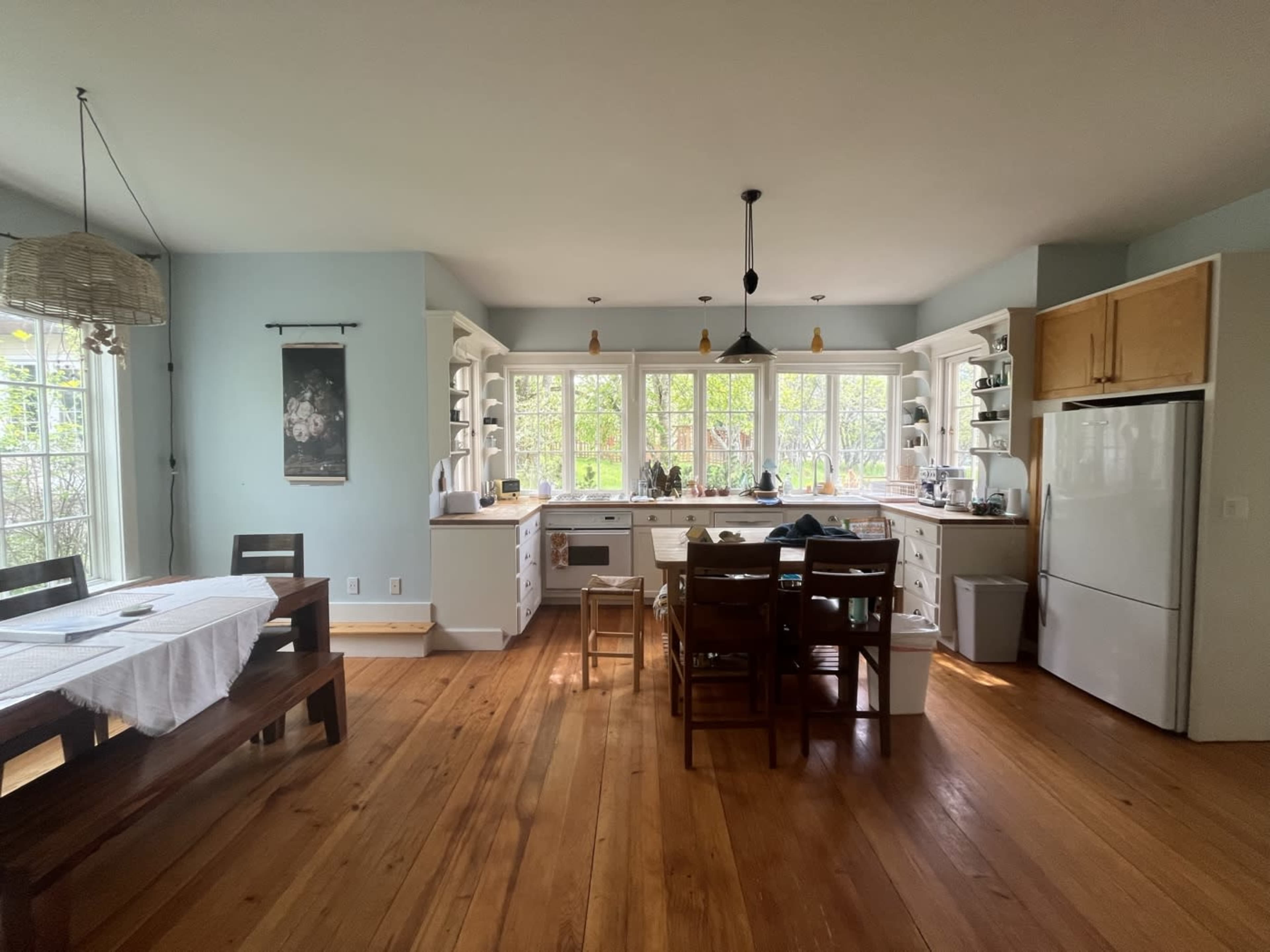 The image shows a bright kitchen with large windows, wooden flooring, and a dining area with a table and chairs.