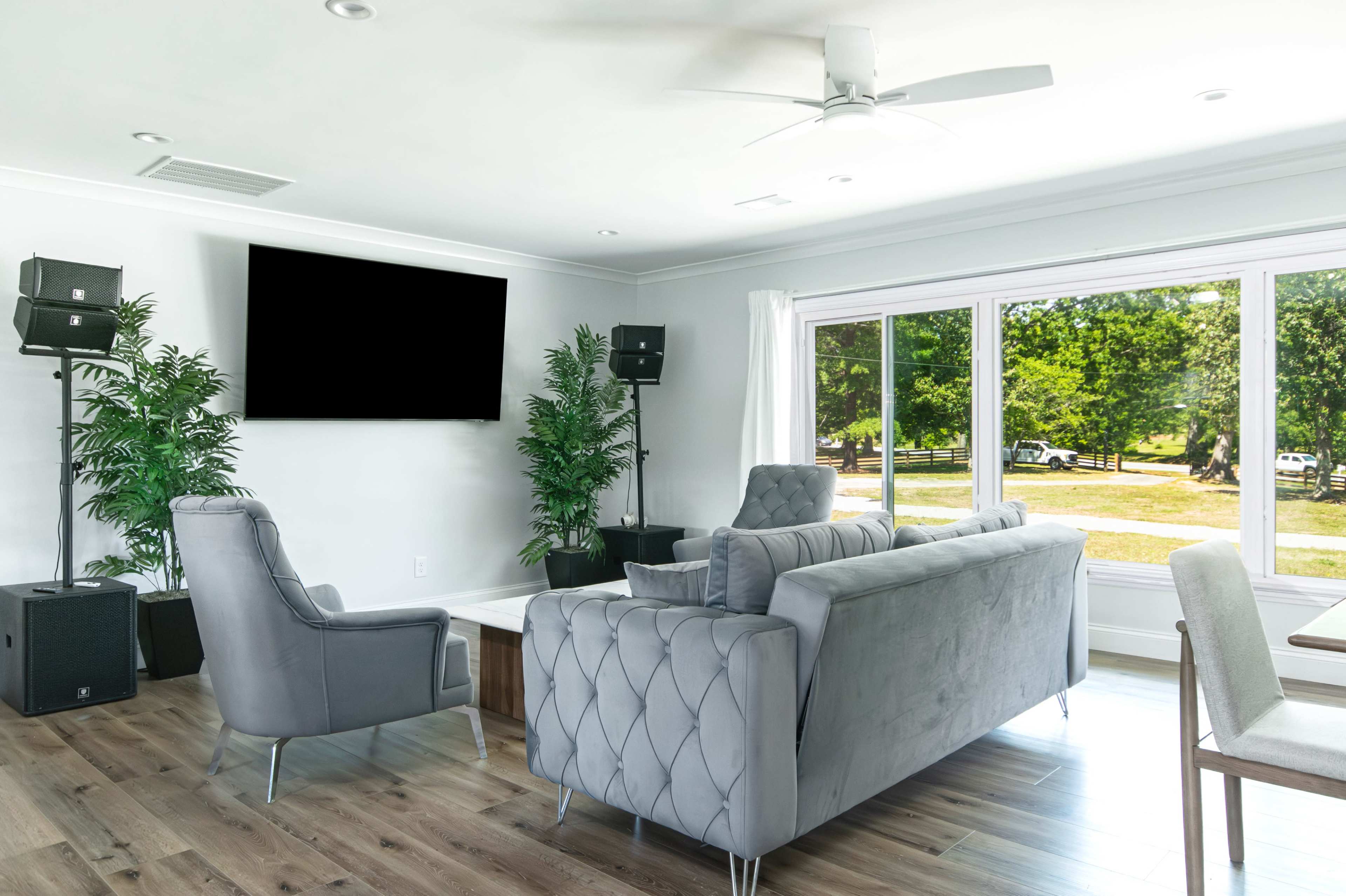 The image shows a modern living room featuring a gray sofa, a single chair, large windows overlooking greenery, and speakers placed in the corners.