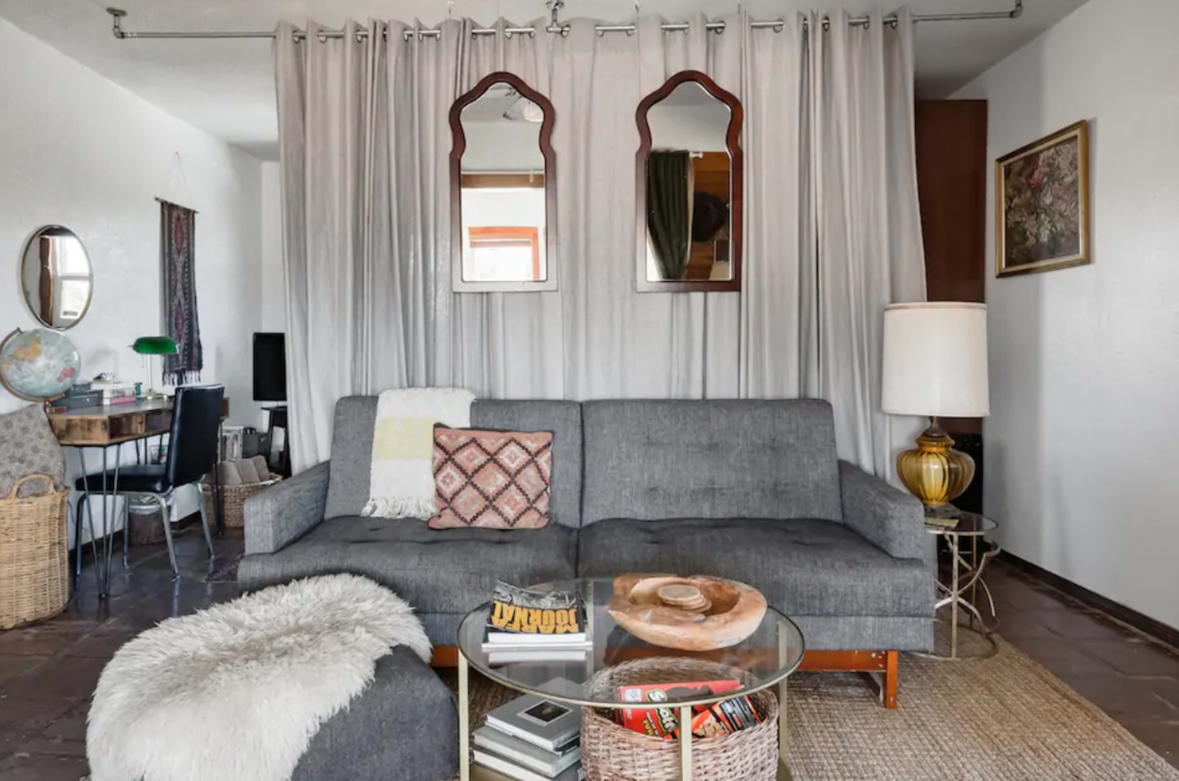 A living room featuring a gray sofa, decorative pillows, a glass coffee table with stacked books, a floor lamp, and curtains draping behind the couch.