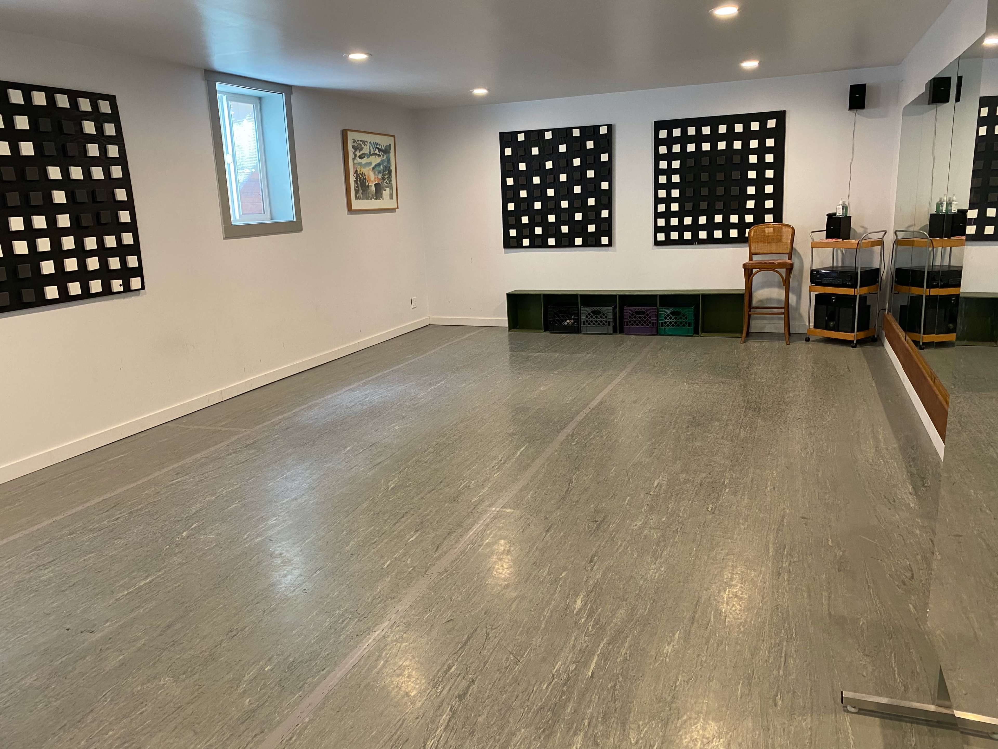 A spacious, well-lit room with green flooring, walls featuring black grid artwork, a mirrored wall, a wooden chair, and storage boxes along one side.
