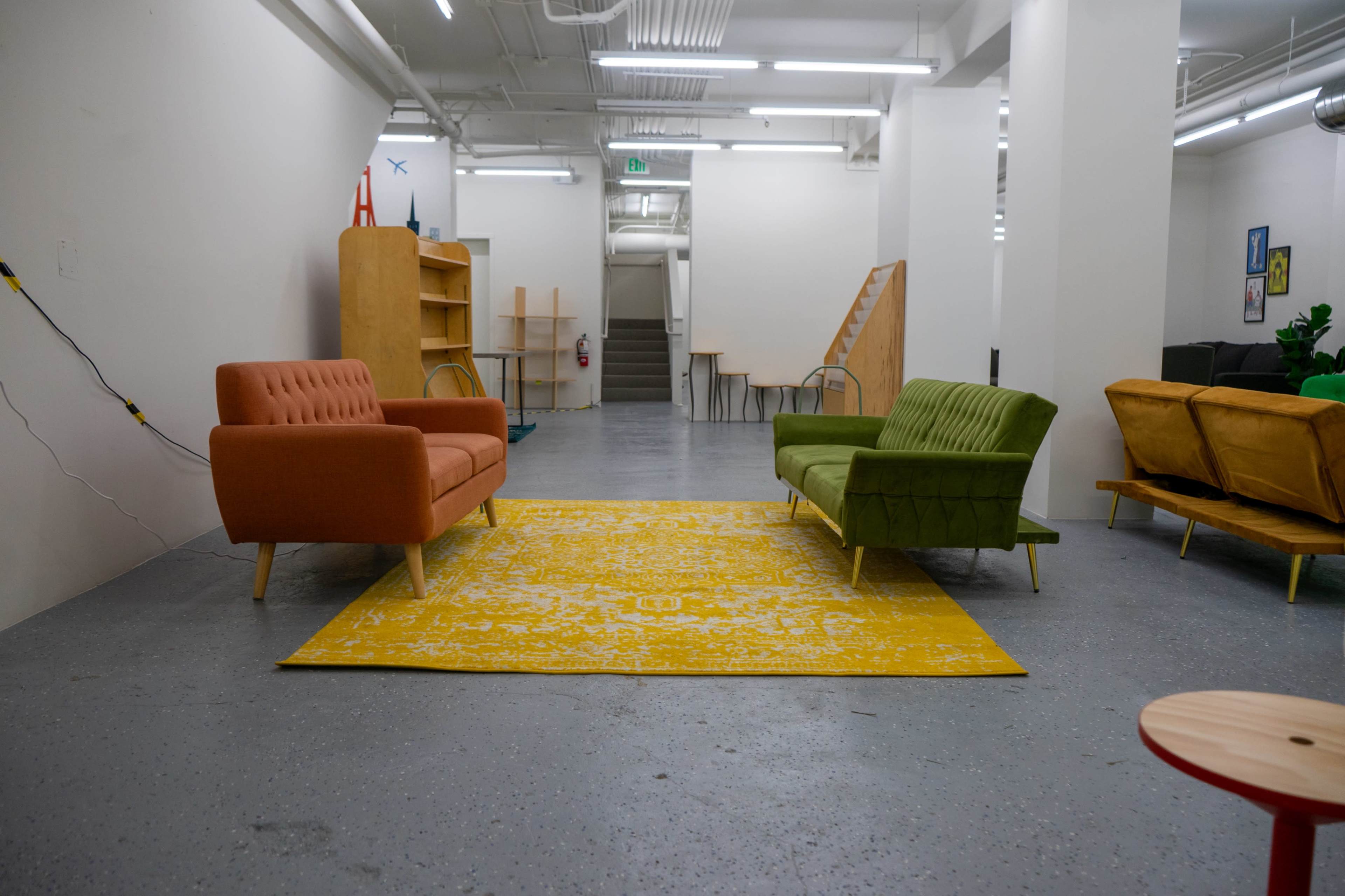 The image shows a spacious, well-lit room with two stylish sofas in orange and green facing each other across a bright yellow rug, and a staircase leading to a higher level in the background.