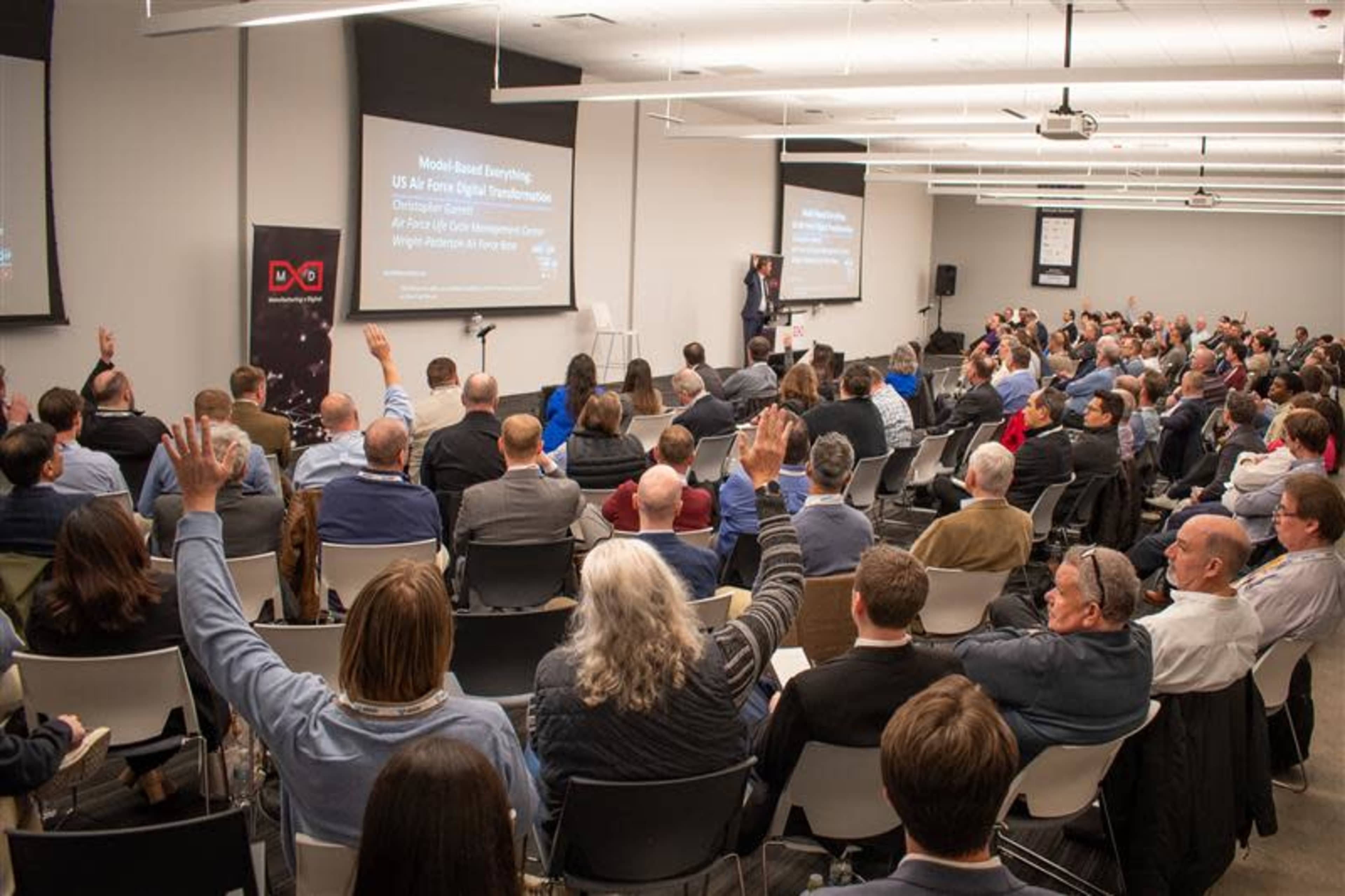 A speaker presents to a large audience in a conference room, with many attendees raising their hands in response to a question.