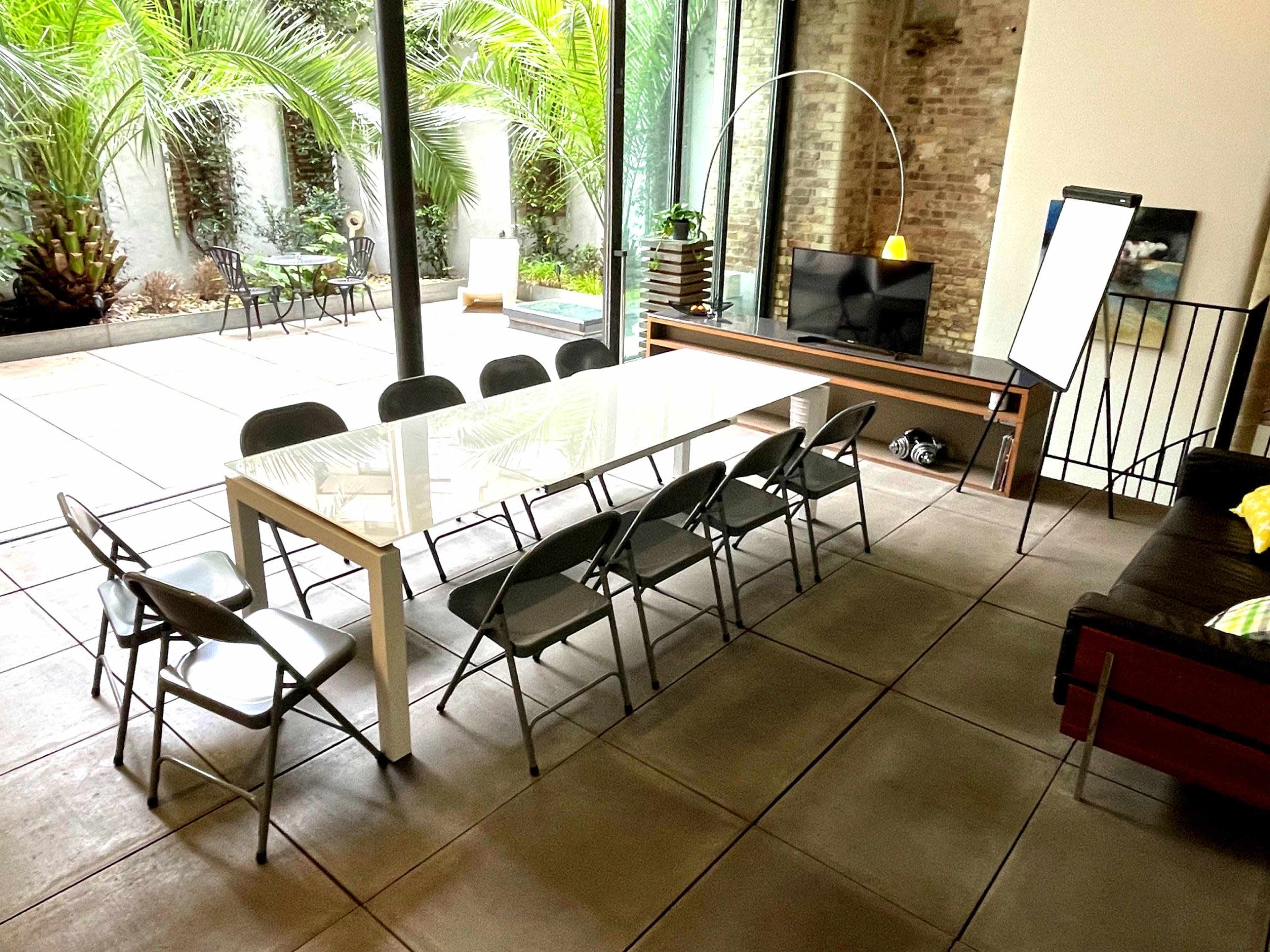 A long white table with eight chairs is set up in a modern space with a view of a lush outdoor patio featuring palm plants.