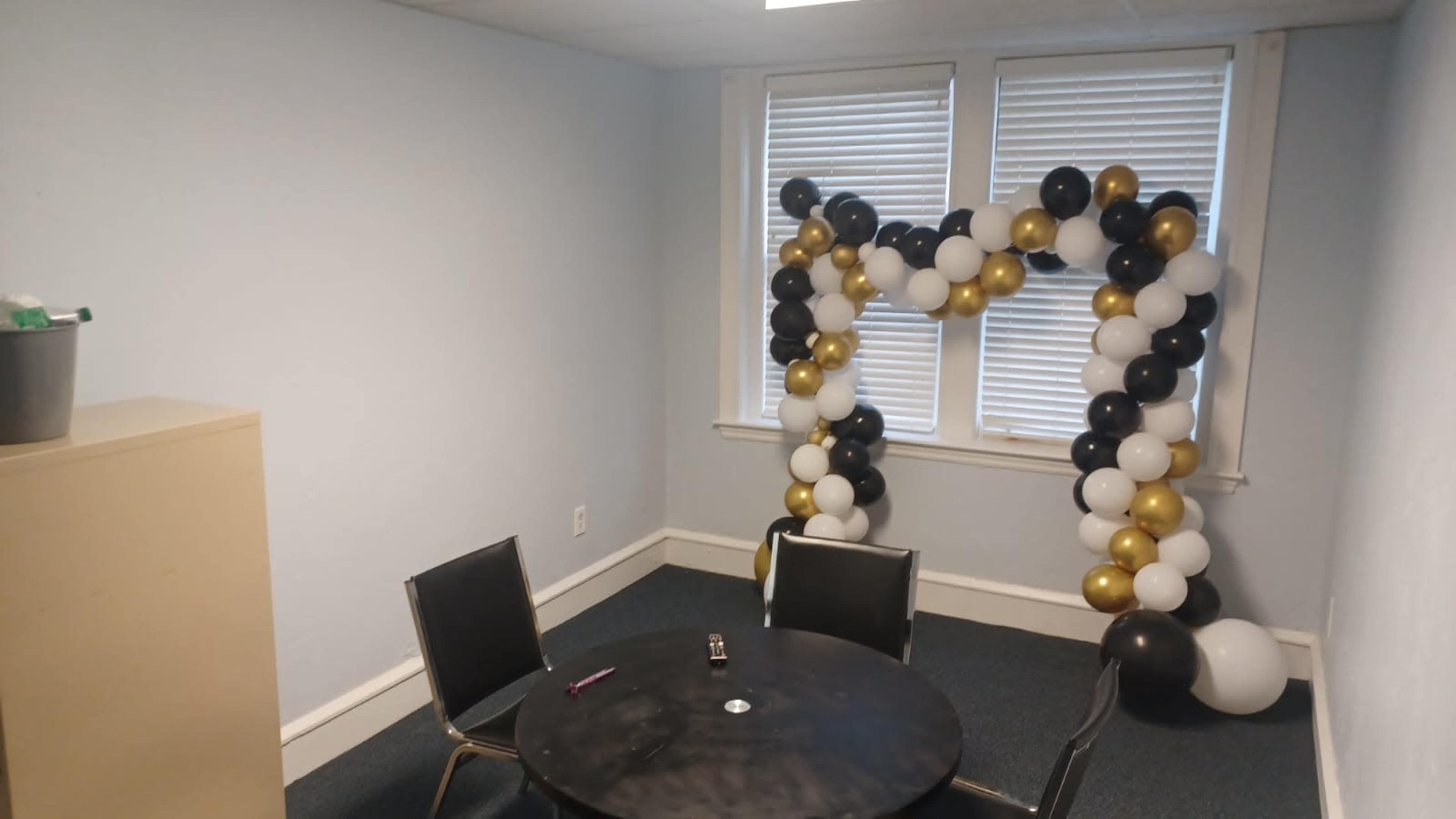 A room features a black circular table with two black chairs and a balloon arch made of black, gold, and white balloons against the wall.
