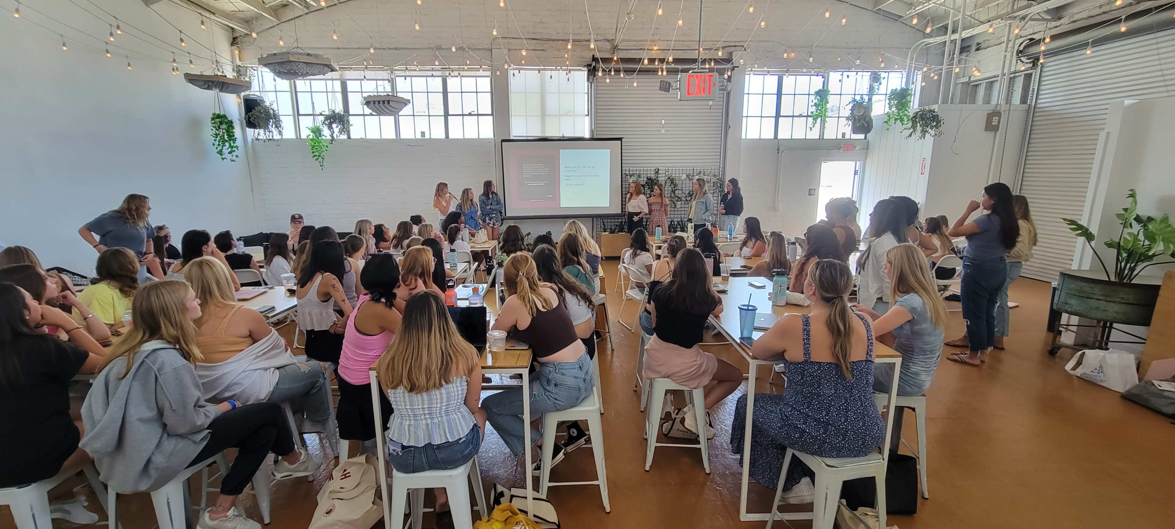A large group of people sits at tables in a bright room, attentively looking towards a screen displaying a presentation.