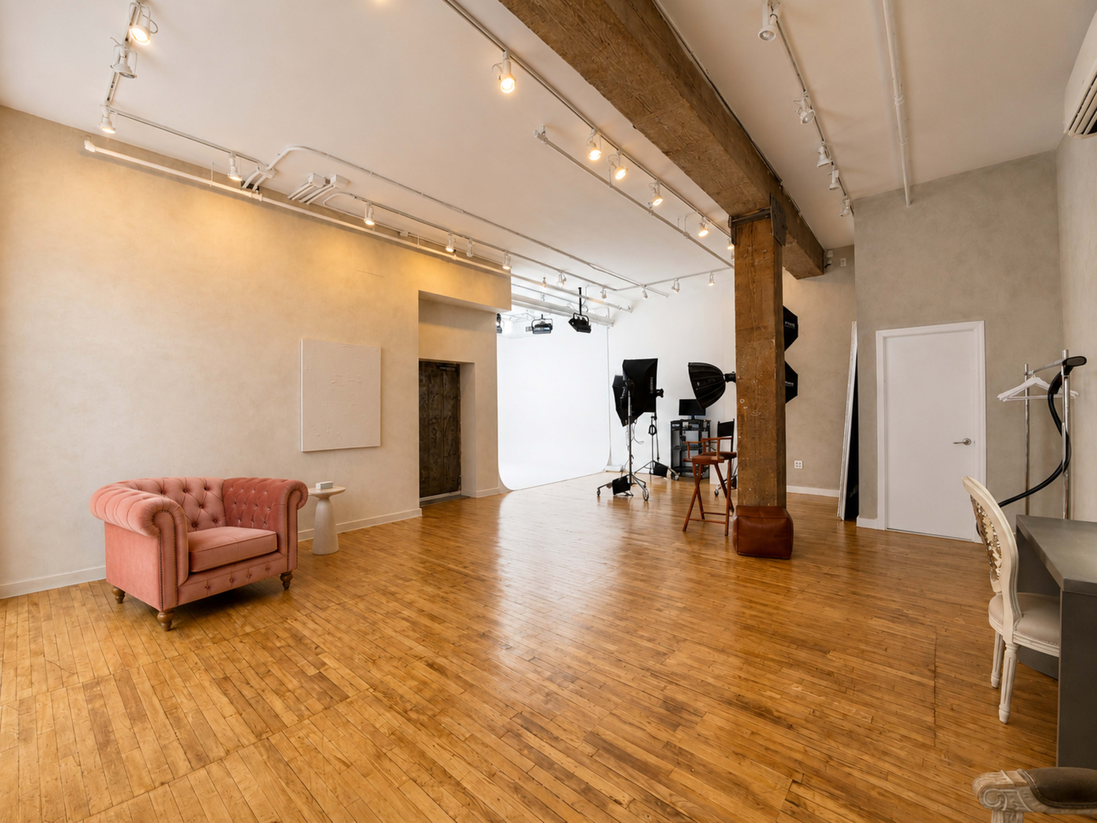 The image shows a spacious photography studio with wooden floors, a large backdrop area, and a pink tufted sofa.