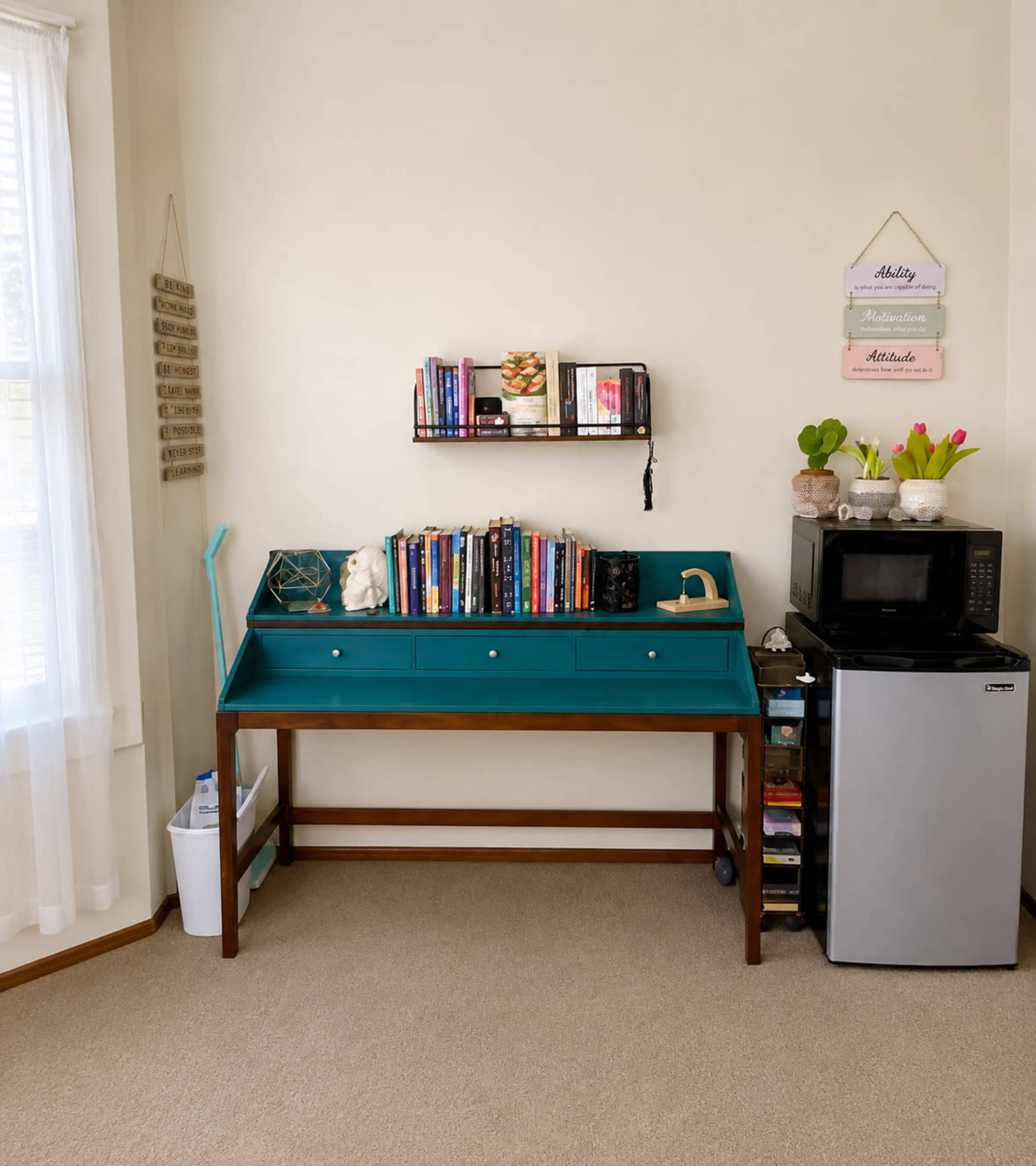 The image shows a teal writing desk with books and a decorative item on top, accompanied by a small bookshelf and a compact microwave beneath it.