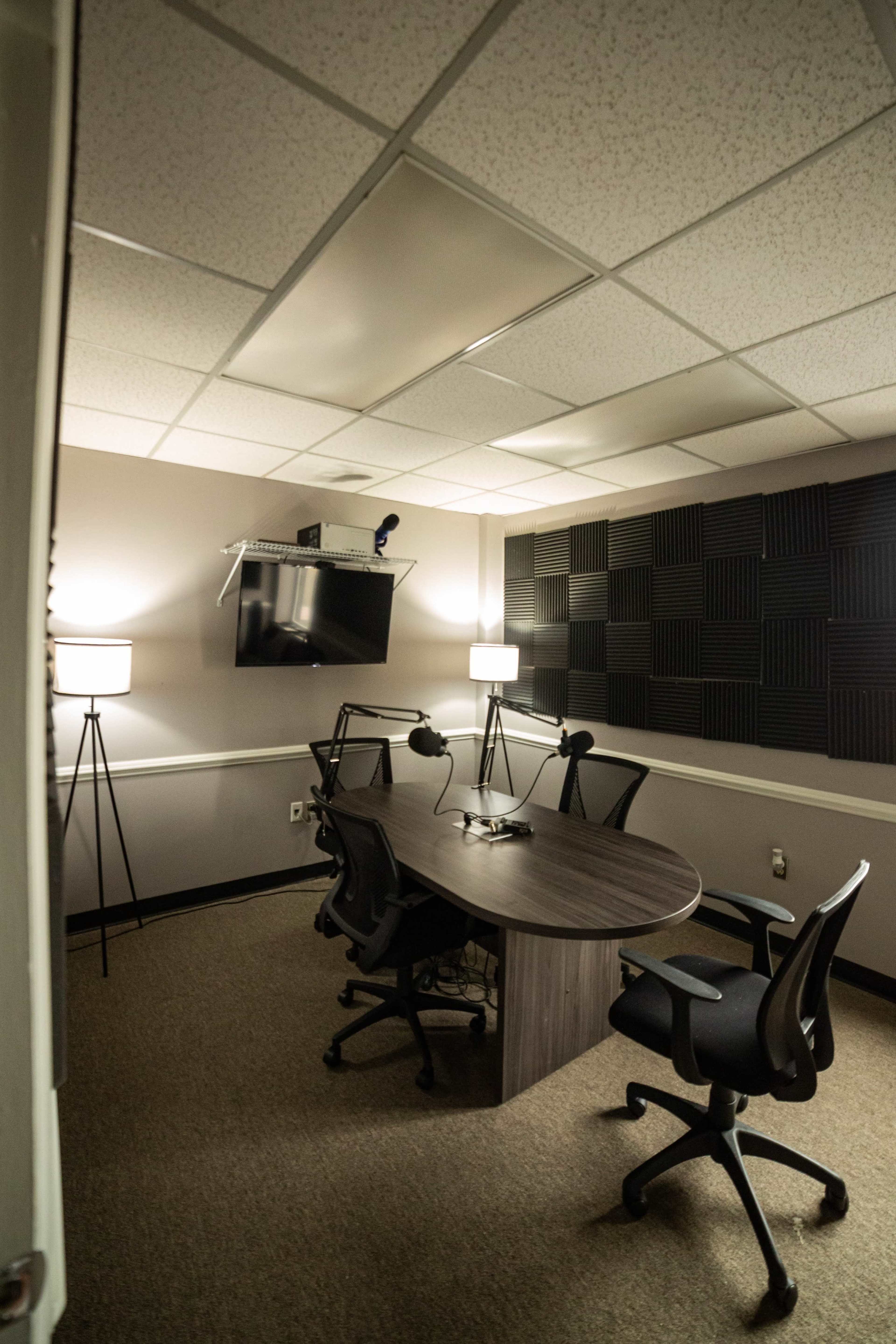 A small recording studio features a large oval table with two chairs, a wall-mounted television, and soundproof paneling.