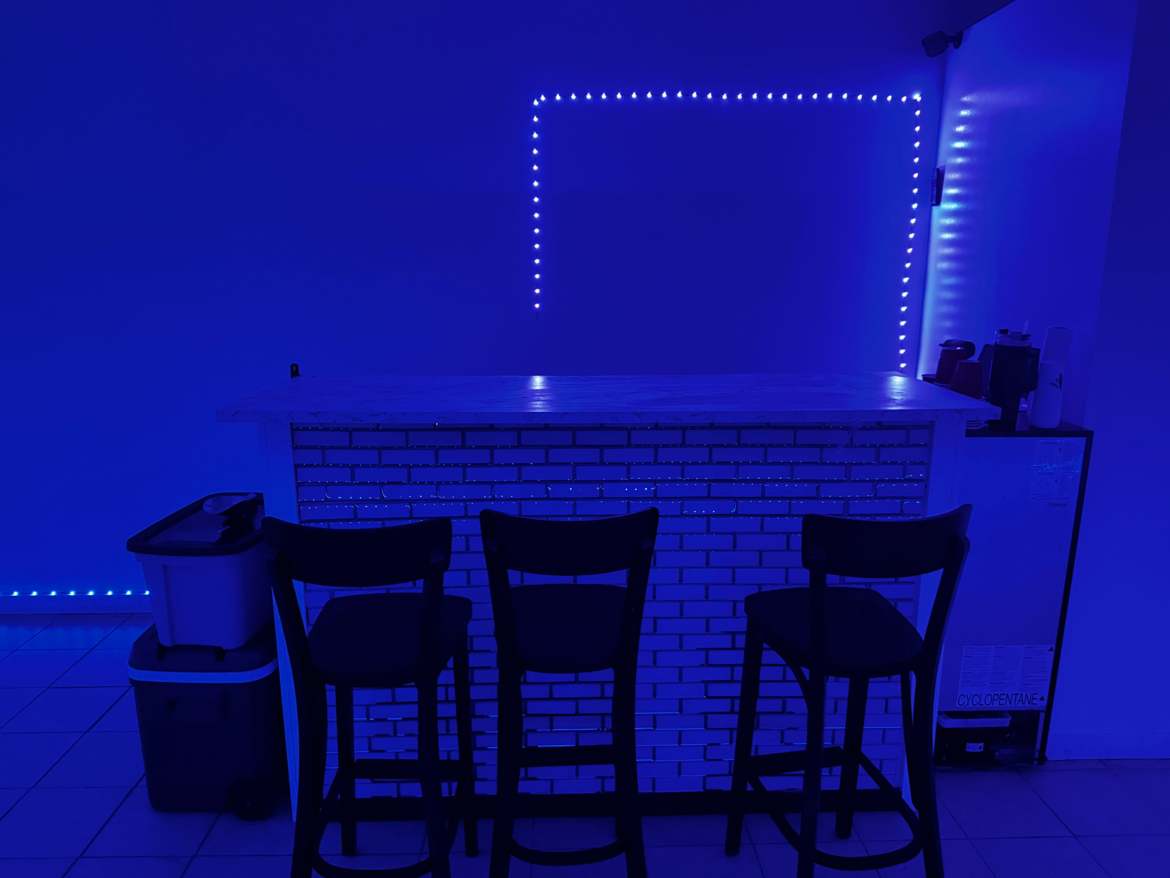 A bar with a white brick front and three black stools is illuminated by blue LED lights.