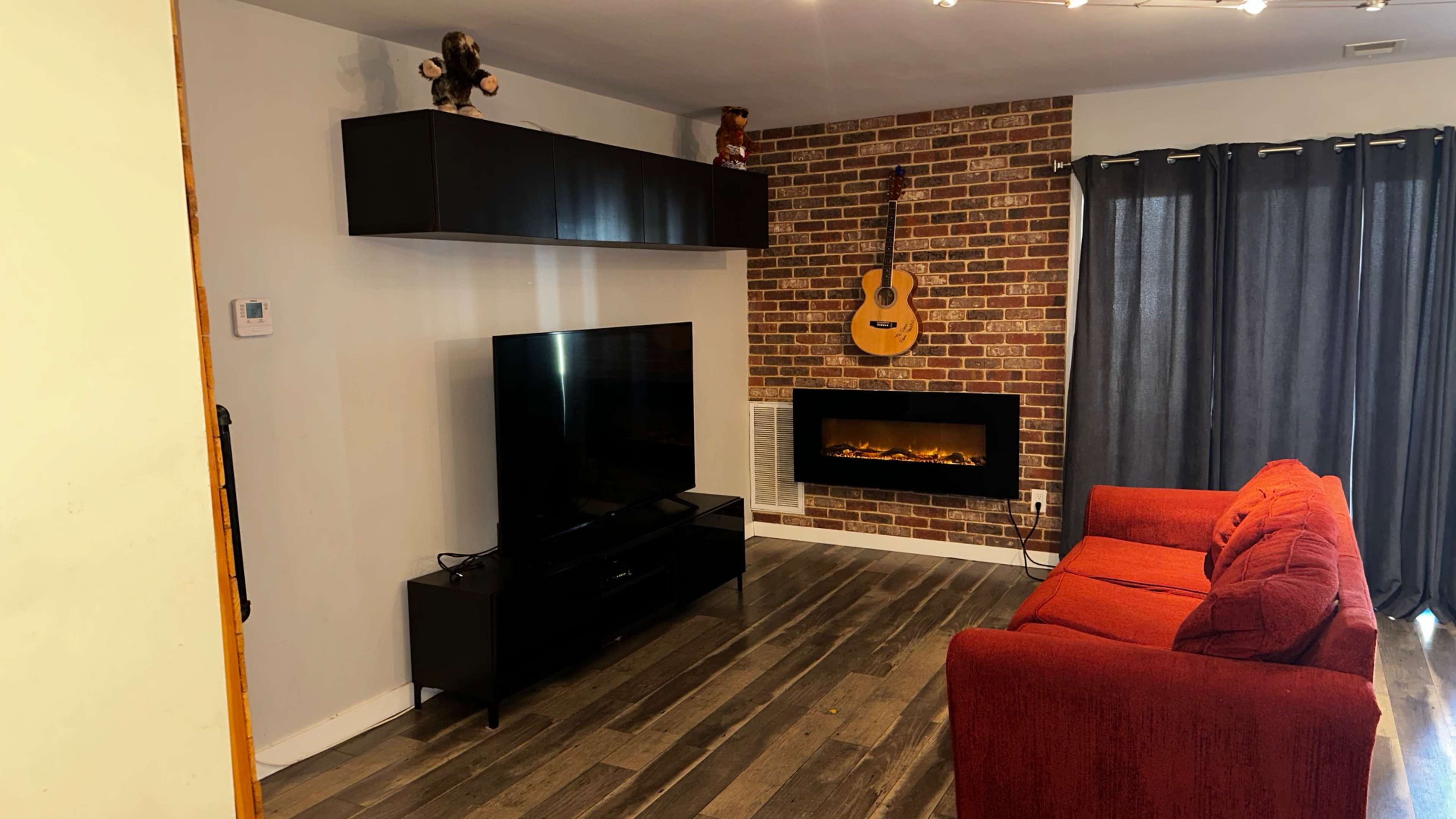 A living room featuring a red sofa, a wall-mounted TV, a brick accent wall with an acoustic guitar, and a faux fireplace below it.