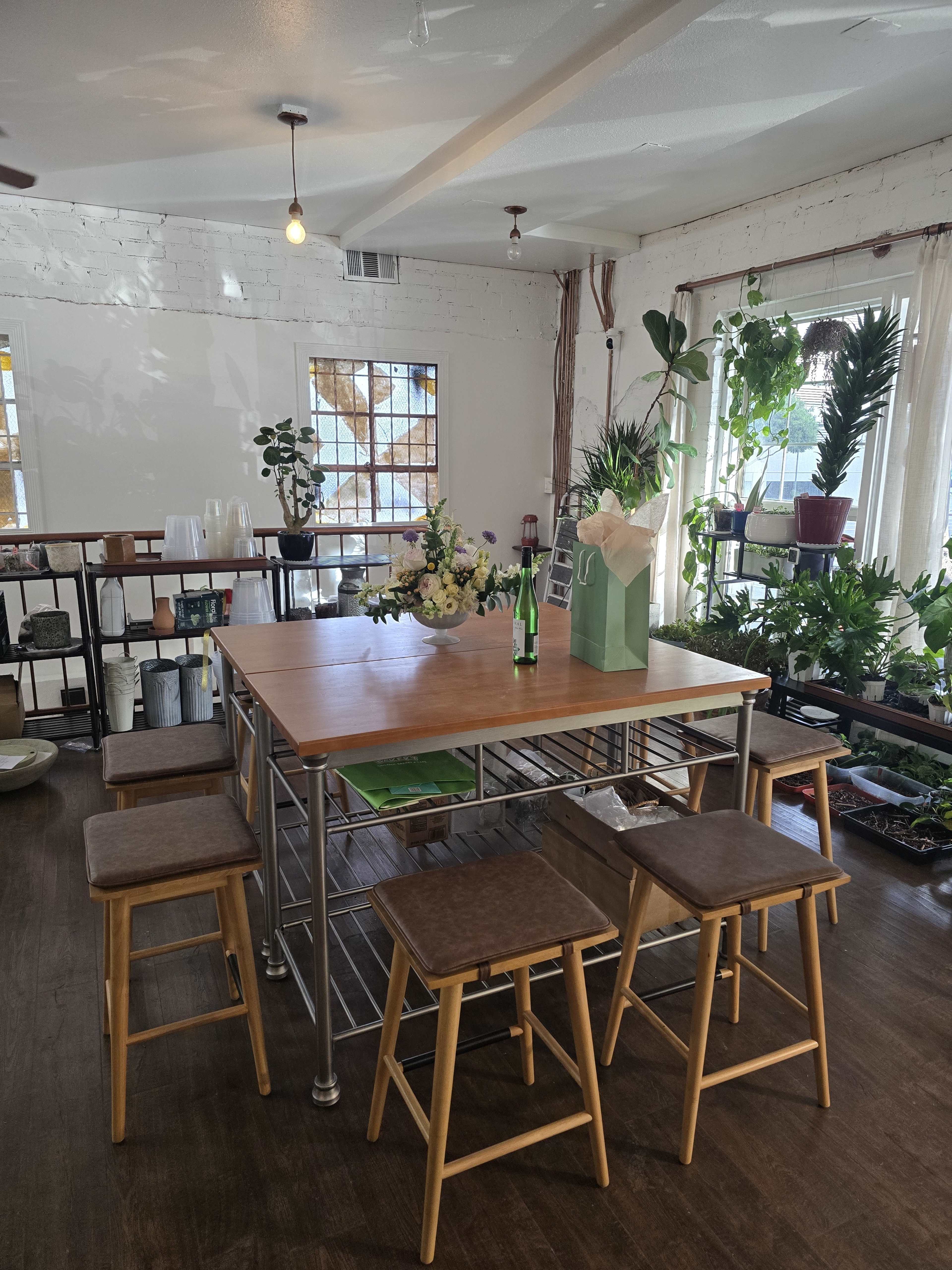 The image shows a spacious room with a wooden table surrounded by stools, featuring various plants and a flower arrangement on the table.