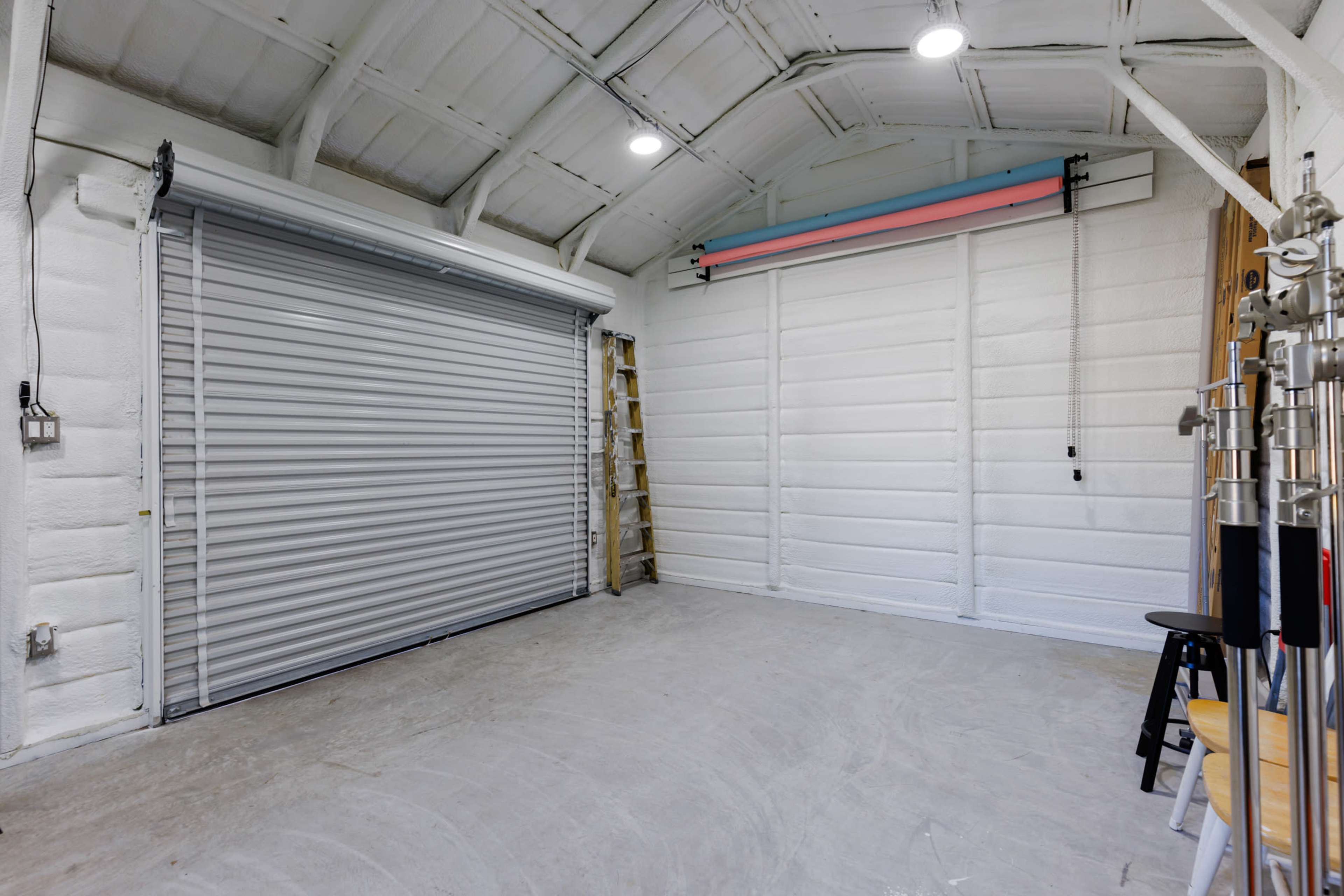 A spacious, empty room with a metal rolling door, a ladder, and a wall-mounted backdrop support.