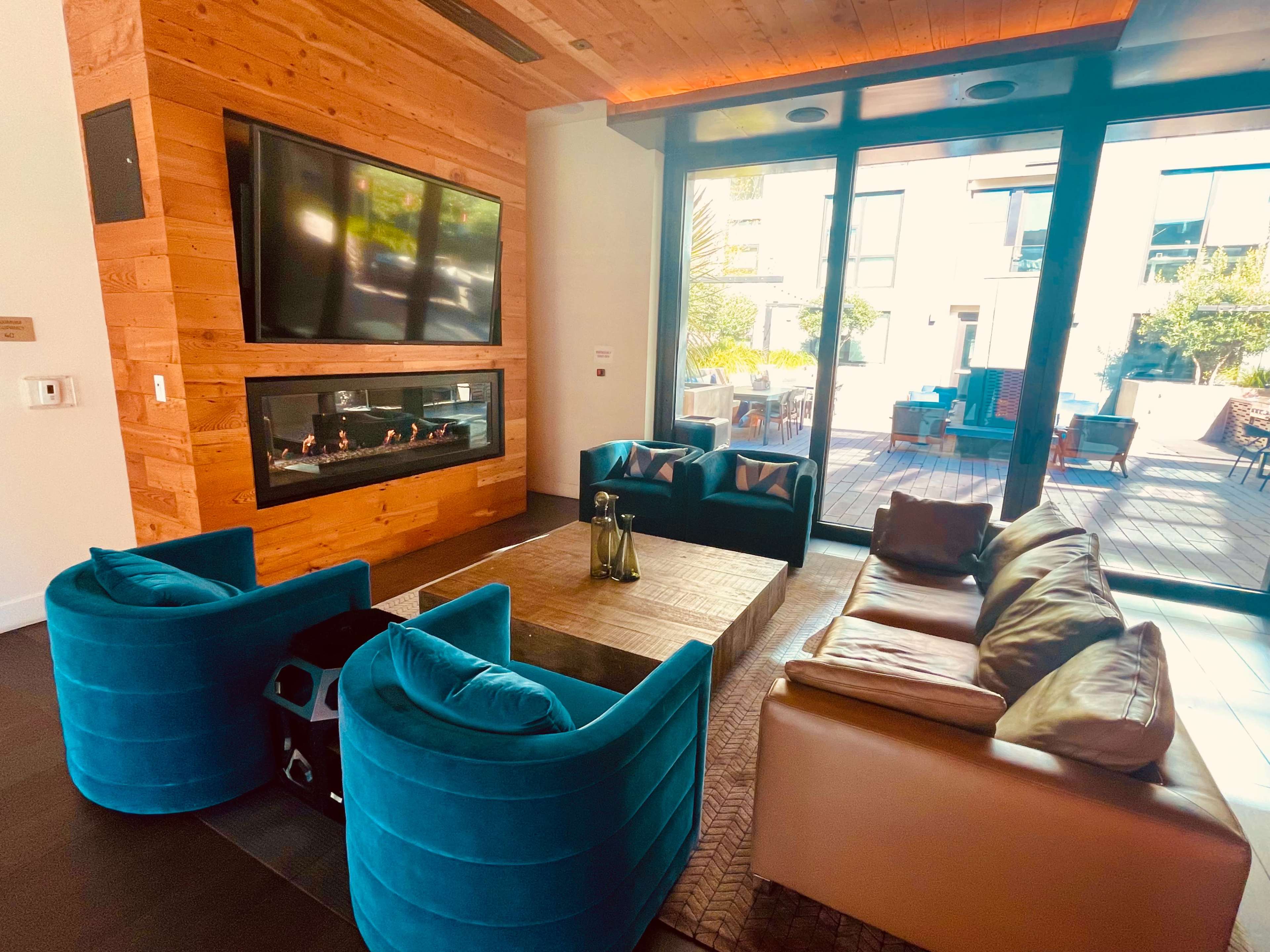A modern living space with a wooden wall, a large window facing an outdoor area, and a seating arrangement that includes blue chairs and a brown sofa surrounding a wooden coffee table.