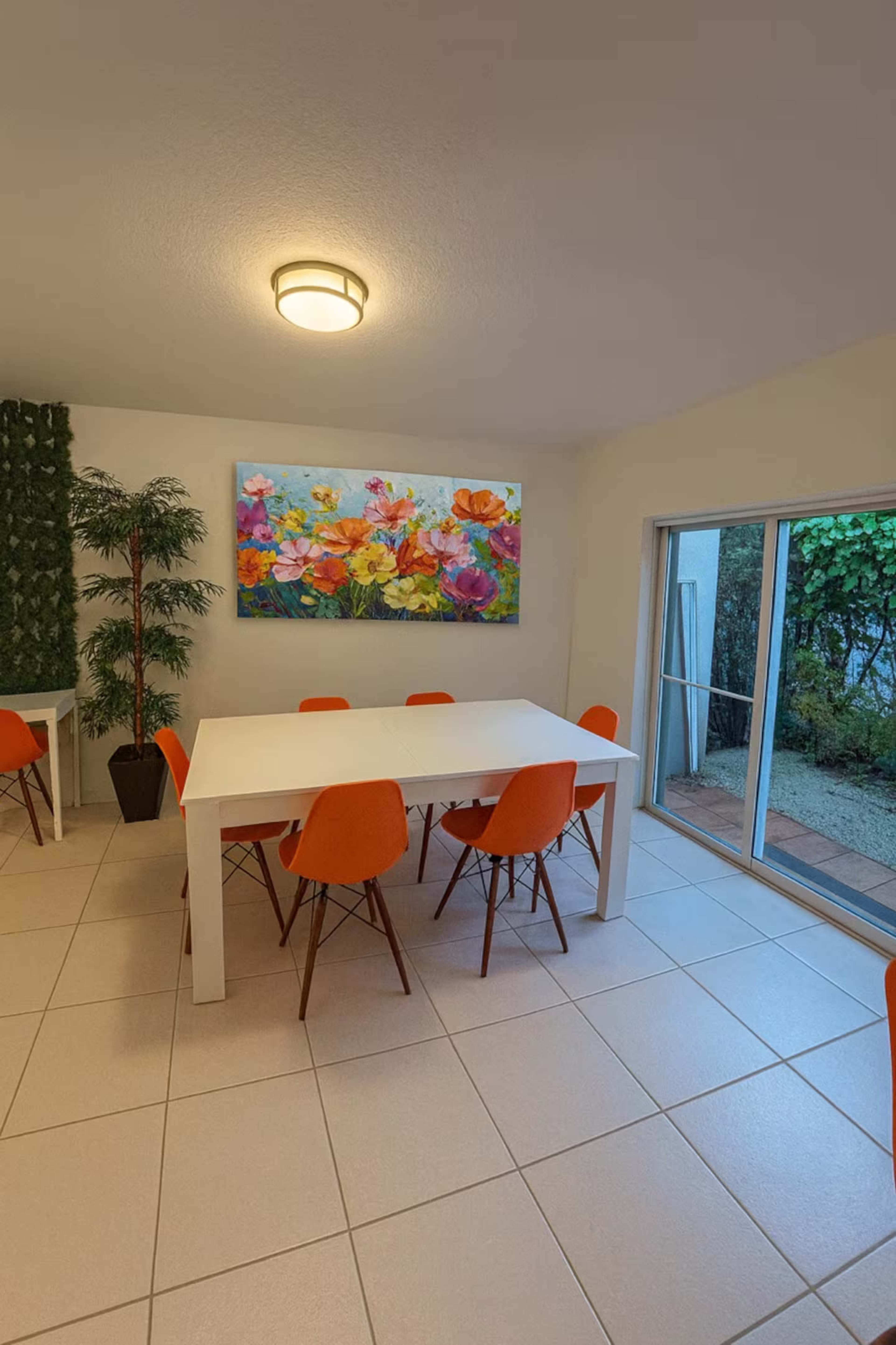 The image shows a dining area with a white table surrounded by orange chairs, complemented by a colorful floral painting on the wall and greenery outside the large sliding doors.