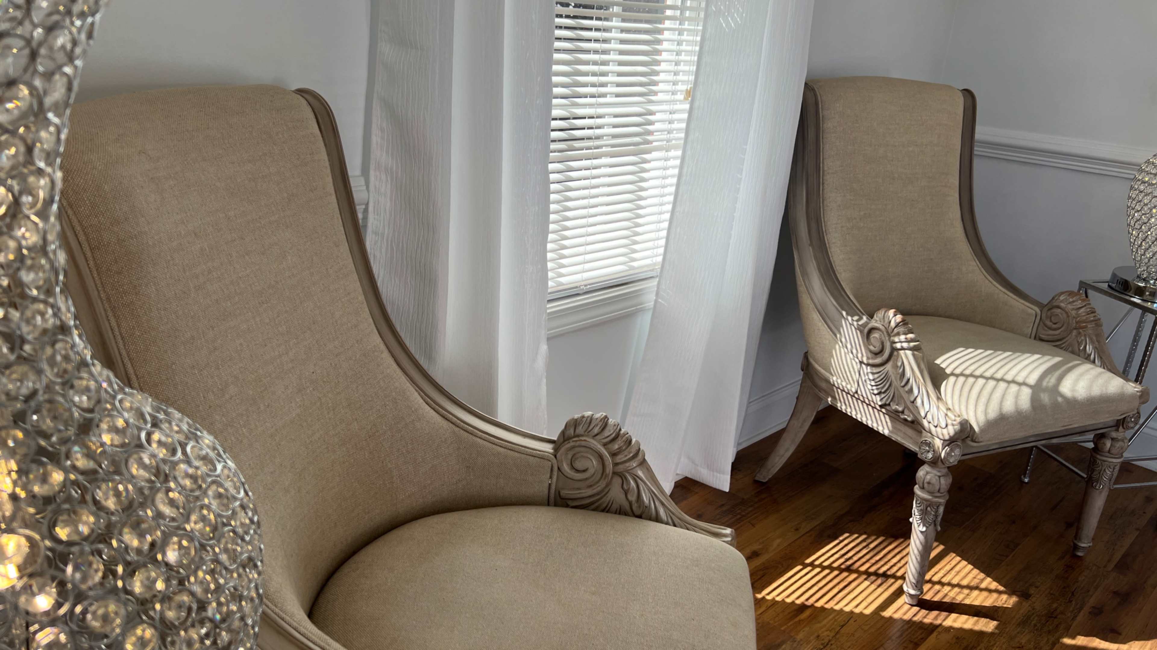 Two upholstered chairs with decorative armrests are positioned near a window, accompanied by a lamp and light streaming through sheer curtains.